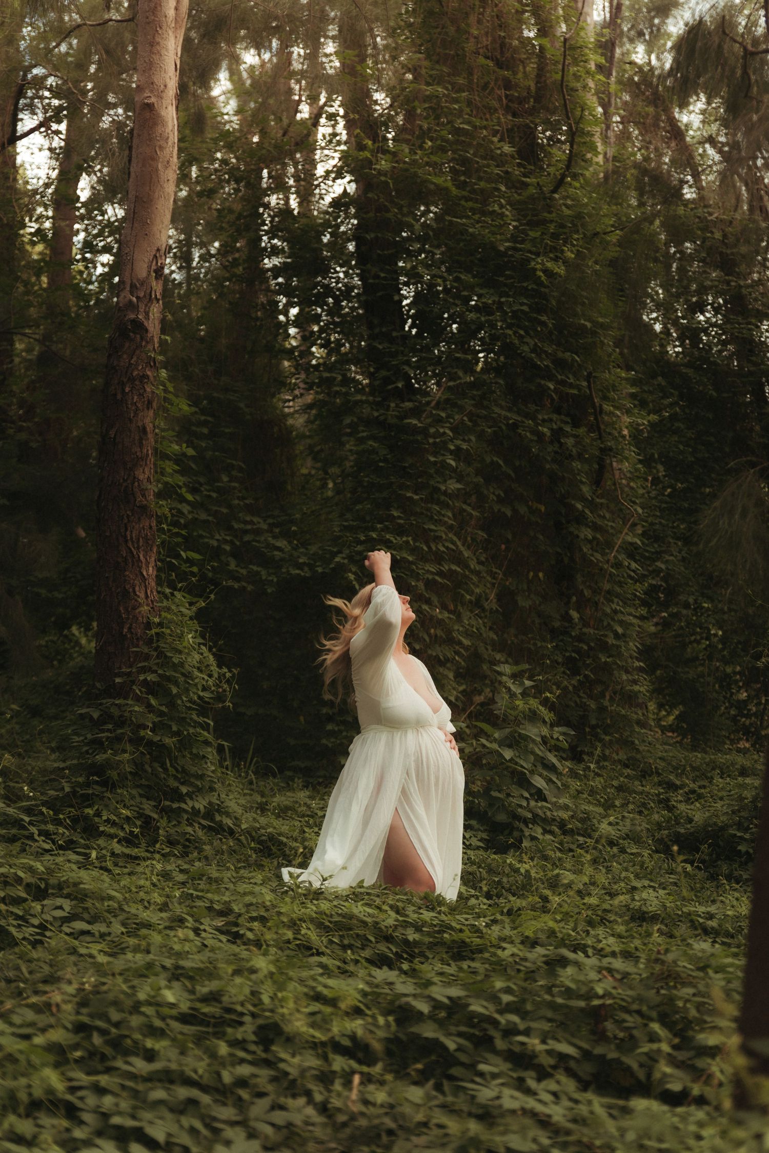 A pregnant woman in a flowing white dress walks through a lush green forest with arms raised.