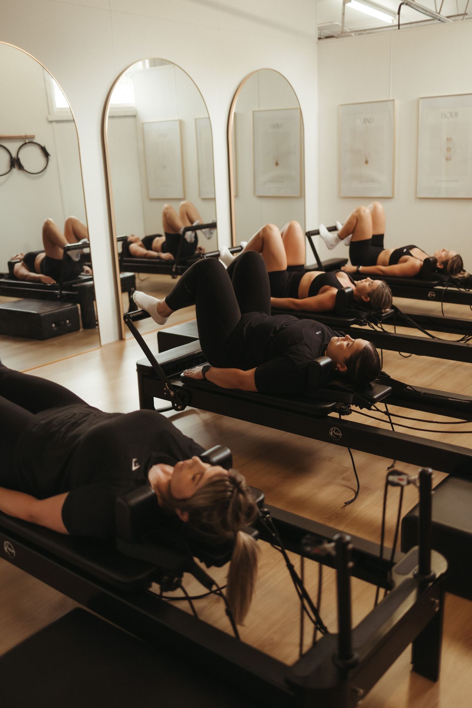 Group of women lying on Pilates reformer machines in elegant studio with arched mirrors and framed artwork.