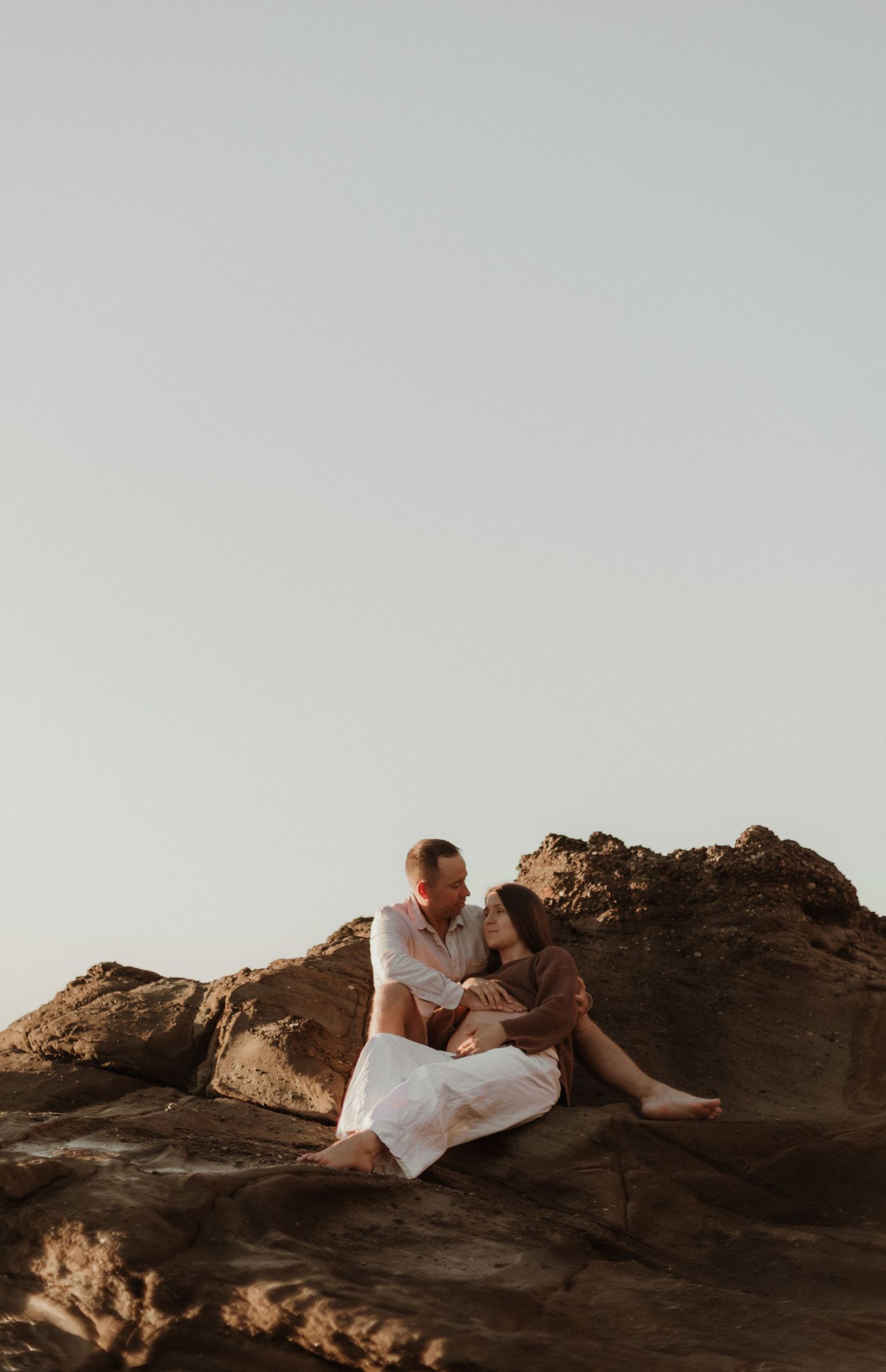 Couple sitting together on rocky outcrop against clear sky at golden hour.