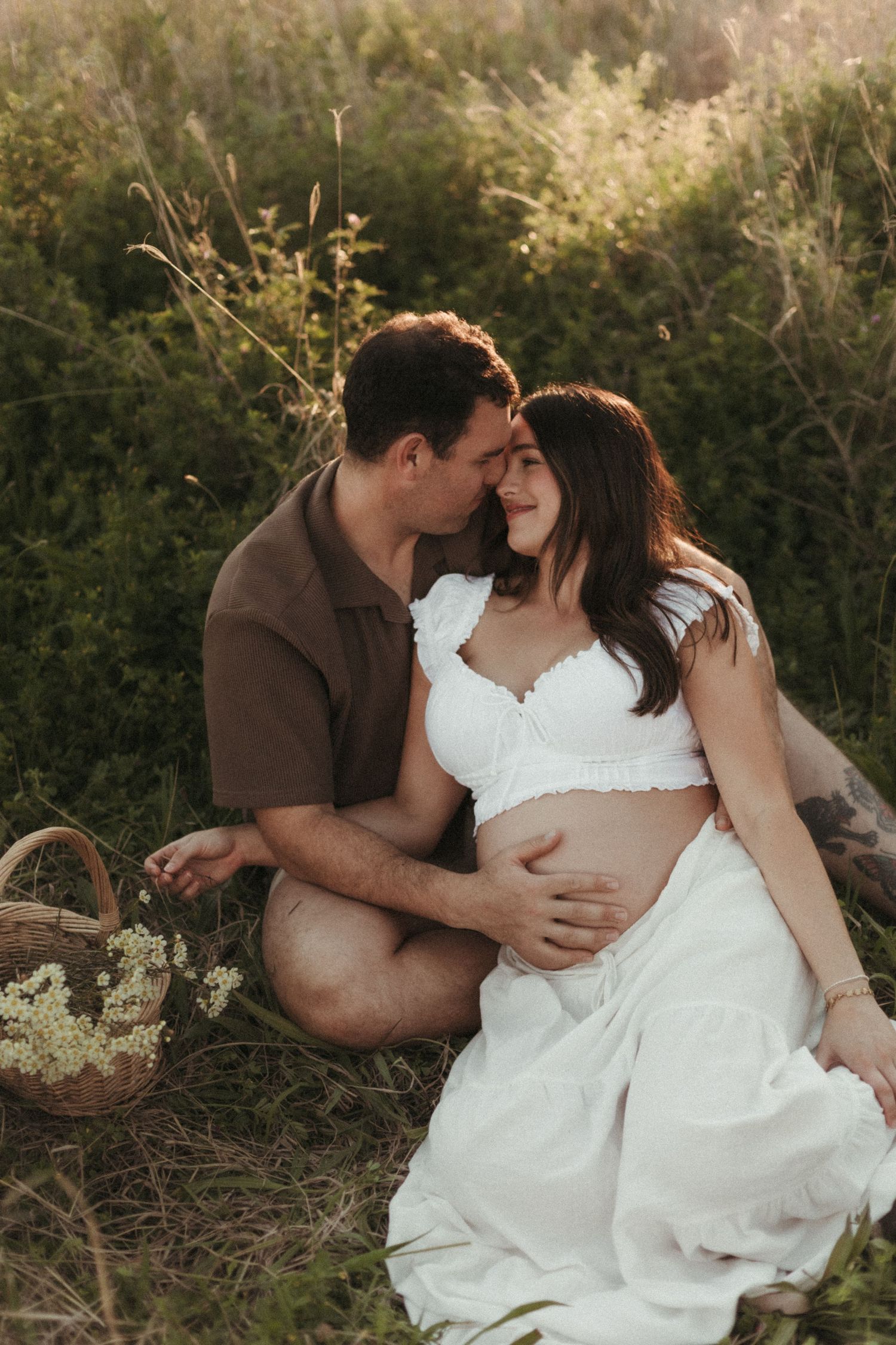 Couple sitting in lush green field, man tenderly embracing pregnant partner at sunset.
