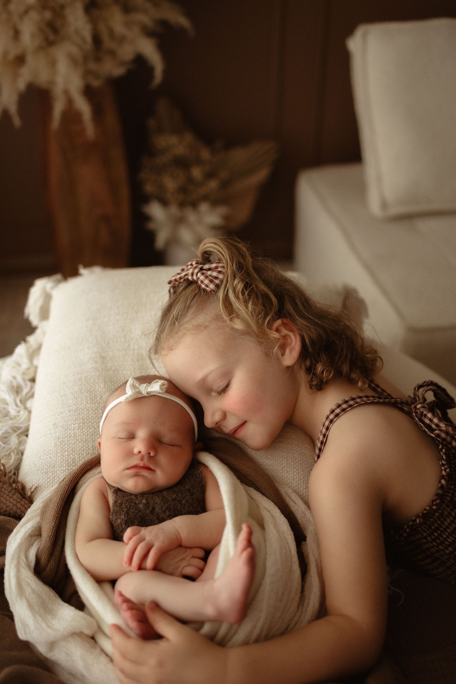 Young girl with curly hair gently rests her cheek on a sleeping newborn baby wrapped in a cozy brown swaddle.