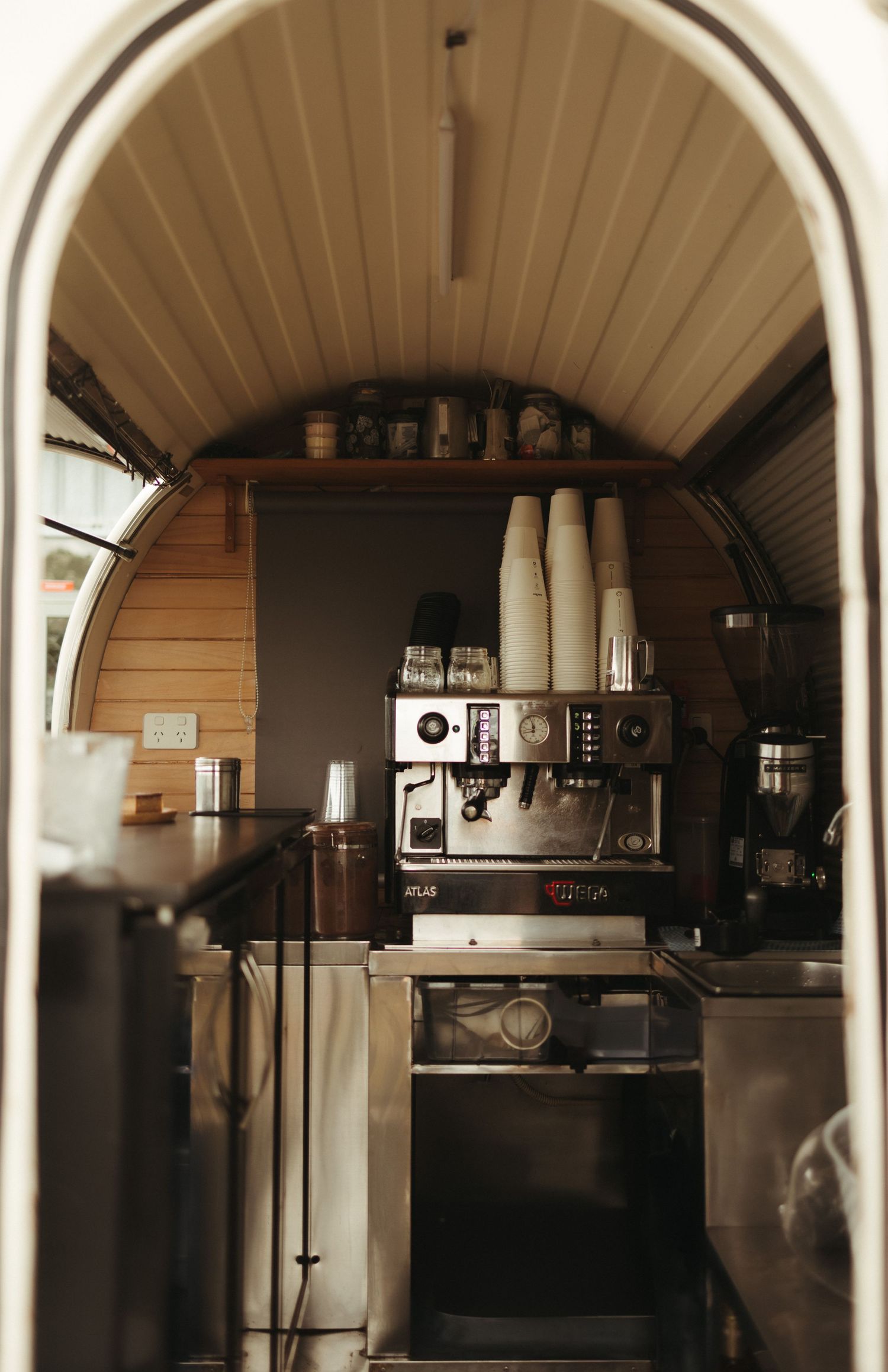 Interior view of a compact coffee van with espresso machine and stacked cups under an arched wooden ceiling.