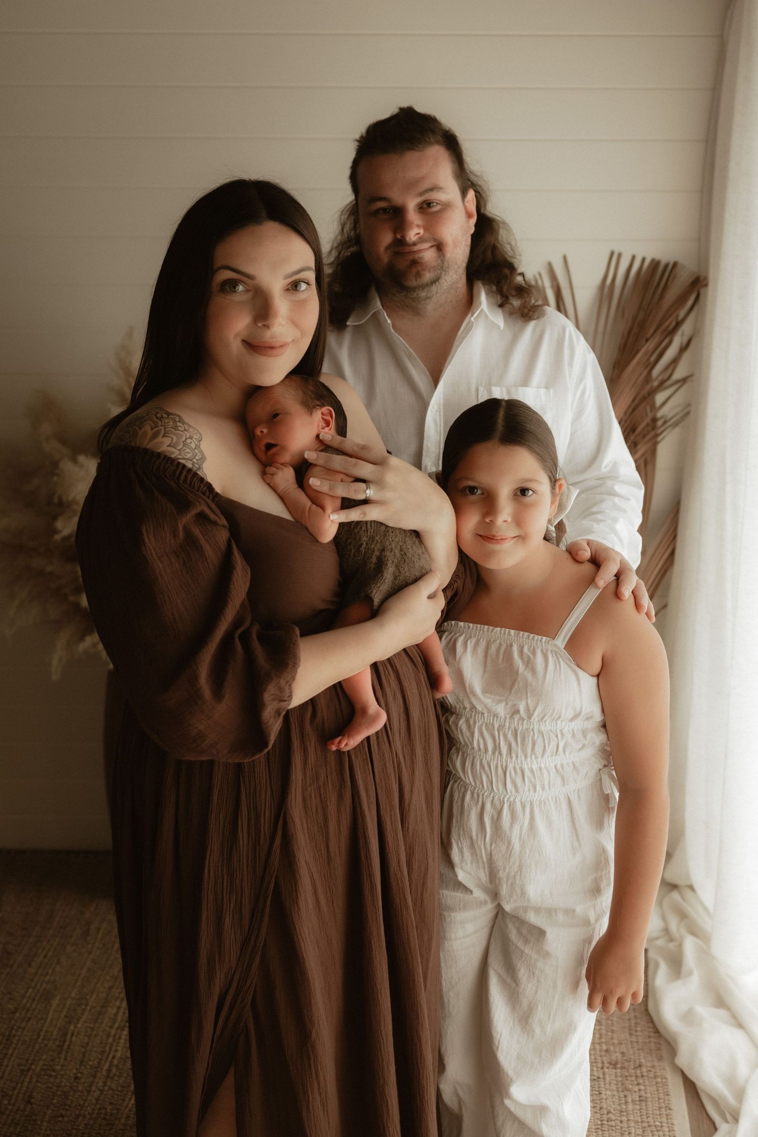 Family portrait with dark-haired mother holding newborn, father and young daughter in boho studio setting.