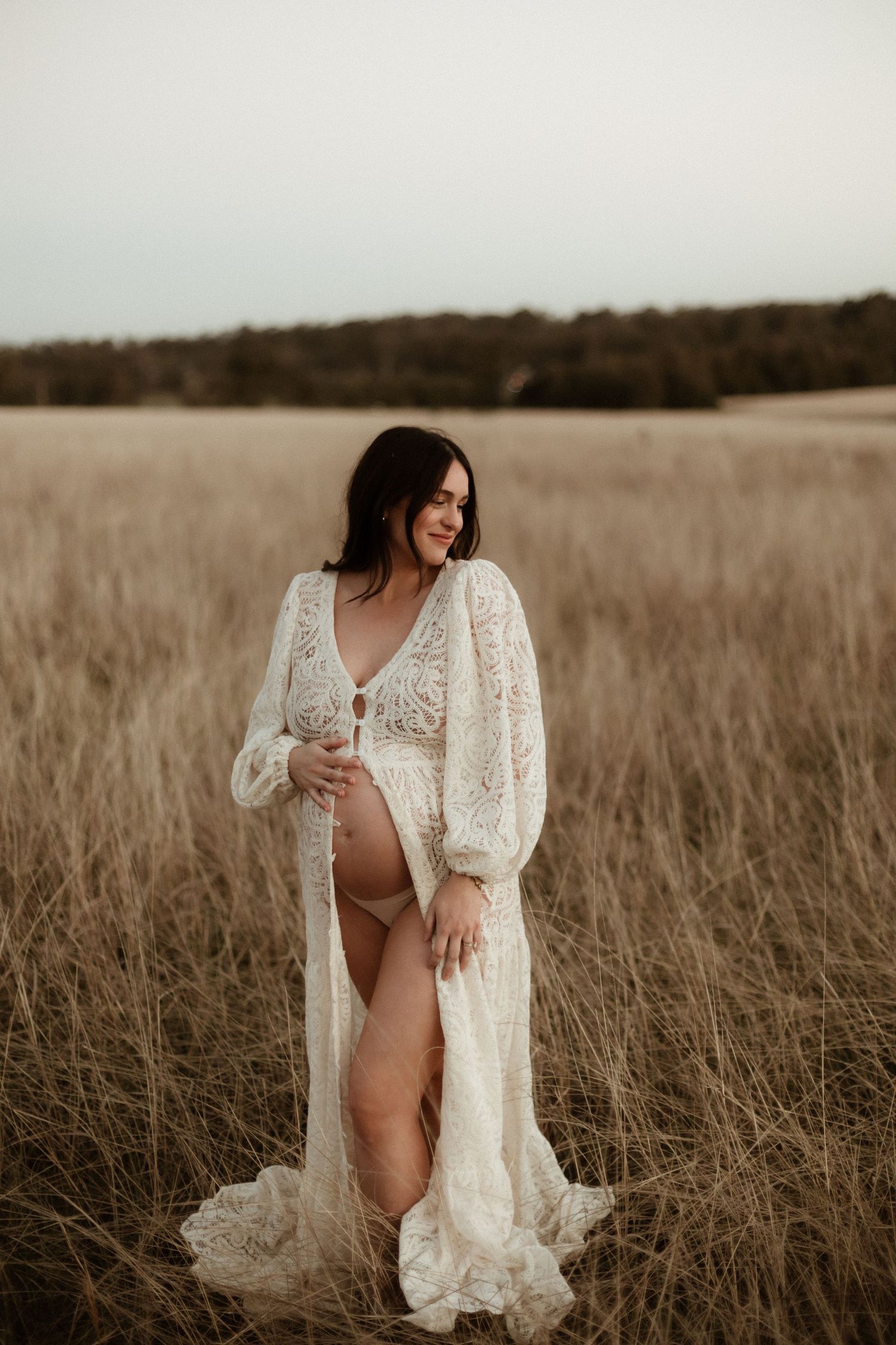 Smiling pregnant woman in white lace open robe posing in golden grass field.