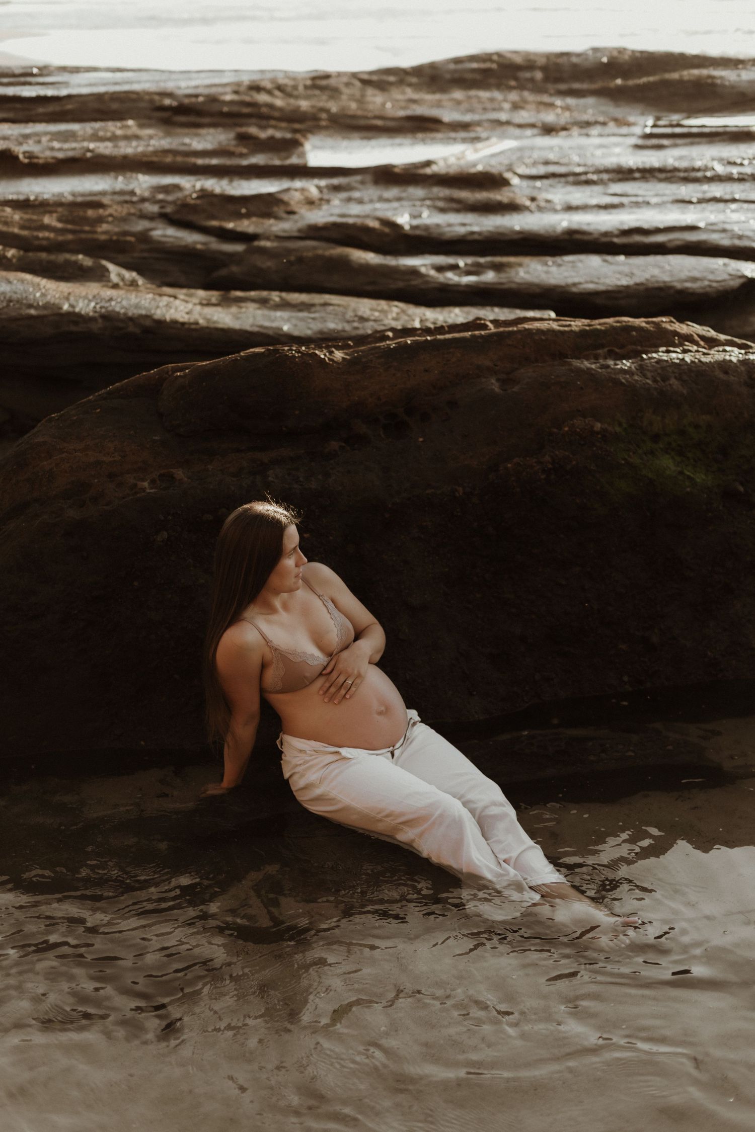 Pregnant woman in bikini top resting against large rocks in shallow water.