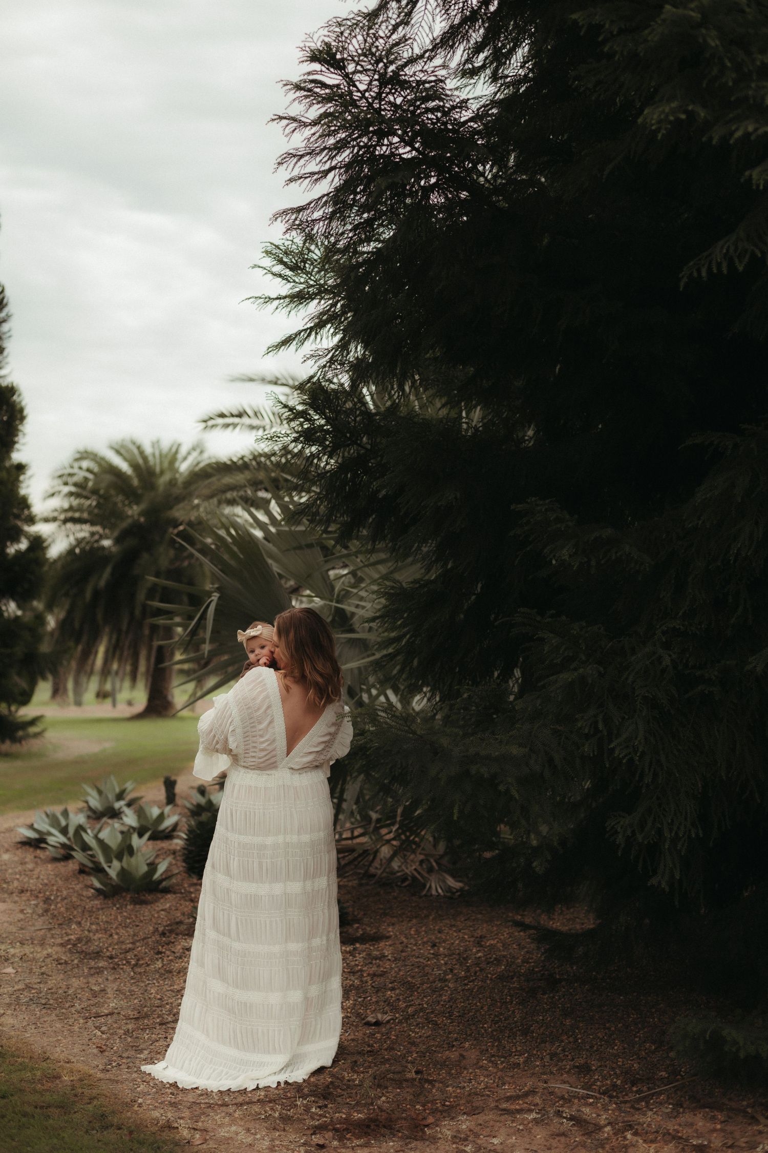 Bride in white boho lace gown standing among lush green trees in garden.