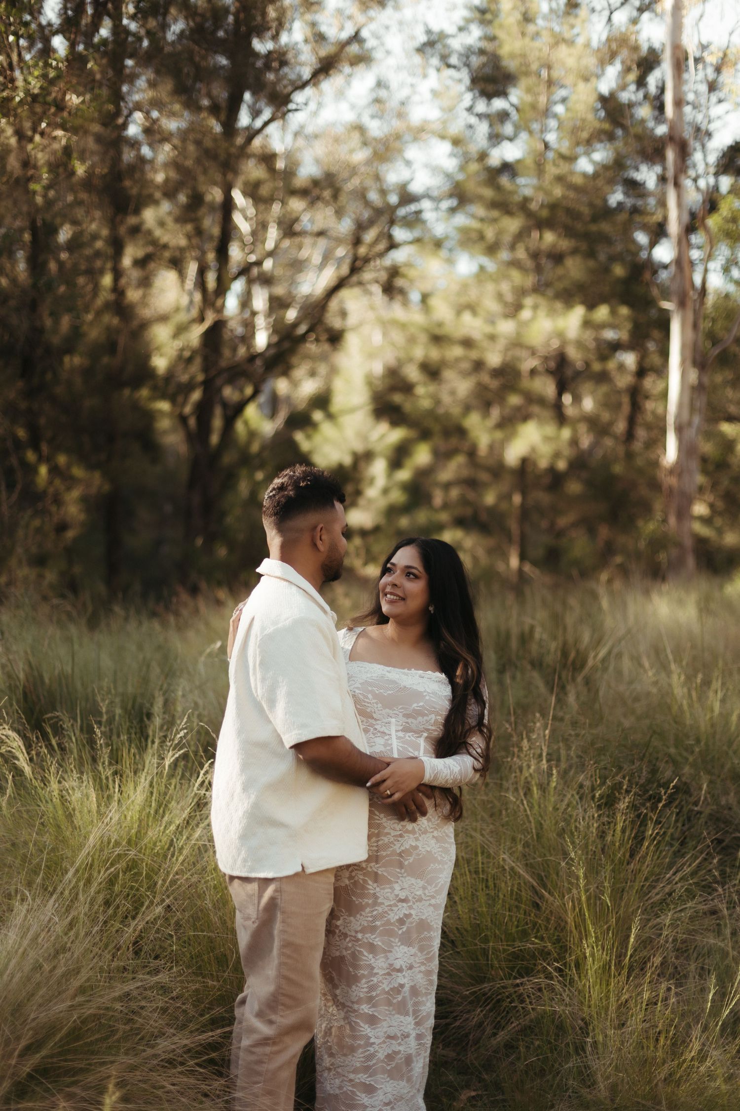Couple embracing in lace wedding dress and white shirt in lush green forest clearing.