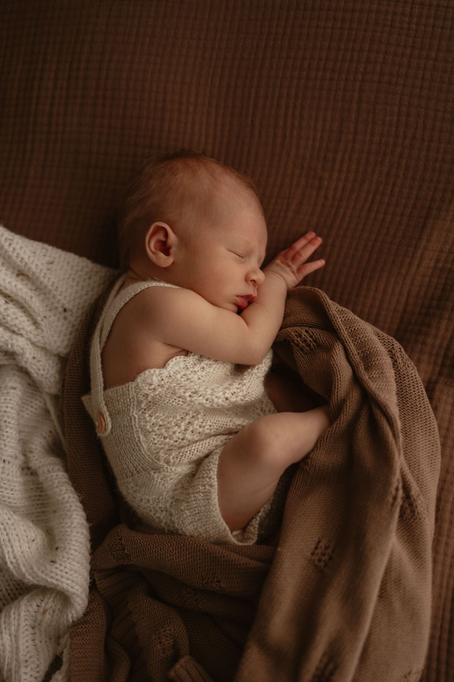 Newborn baby in cream knit romper sleeping peacefully on brown textured blanket with hand near face.