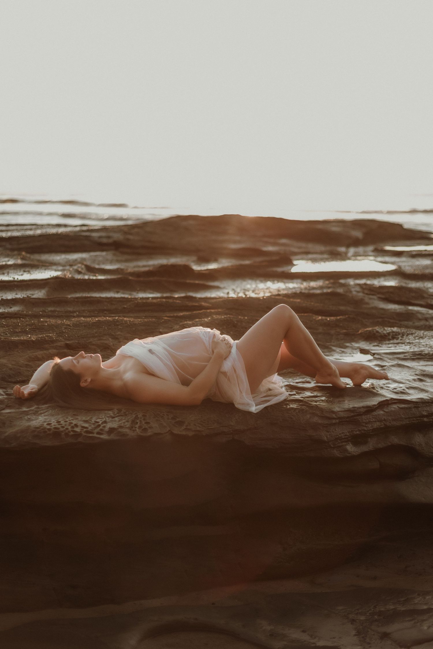 Pregnant woman in white lying on rocky beach surface in warm sunset light.