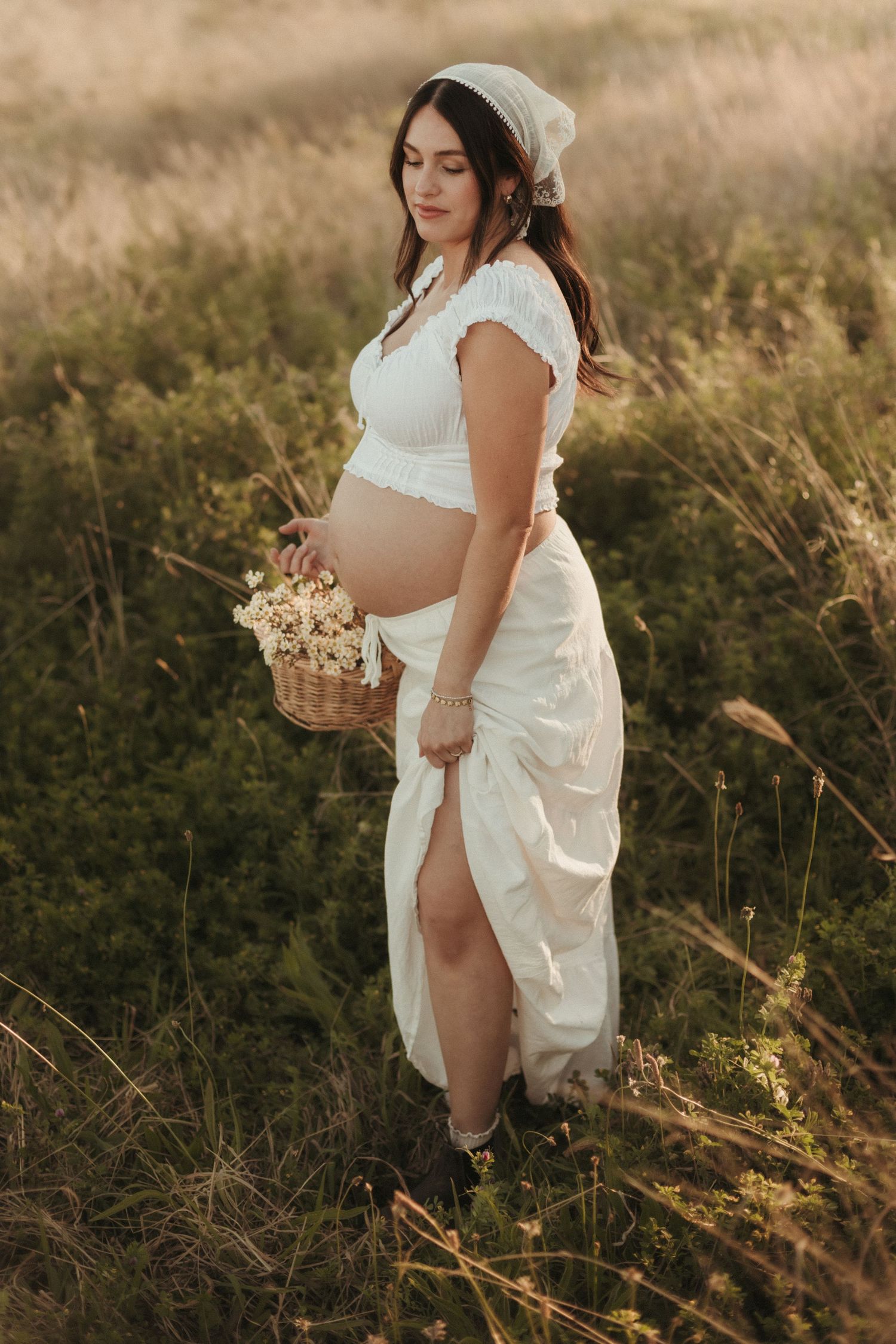 Pregnant woman in white lace outfit holding flower basket in sunlit grassy field.