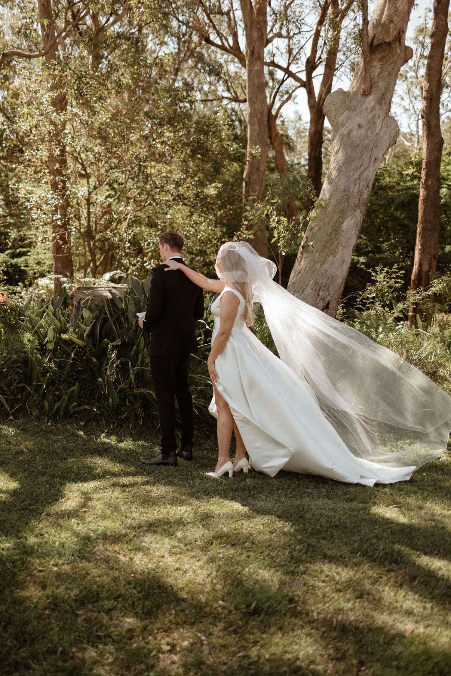 Bride in flowing white gown and veil walks with groom in black tuxedo through lush garden setting.