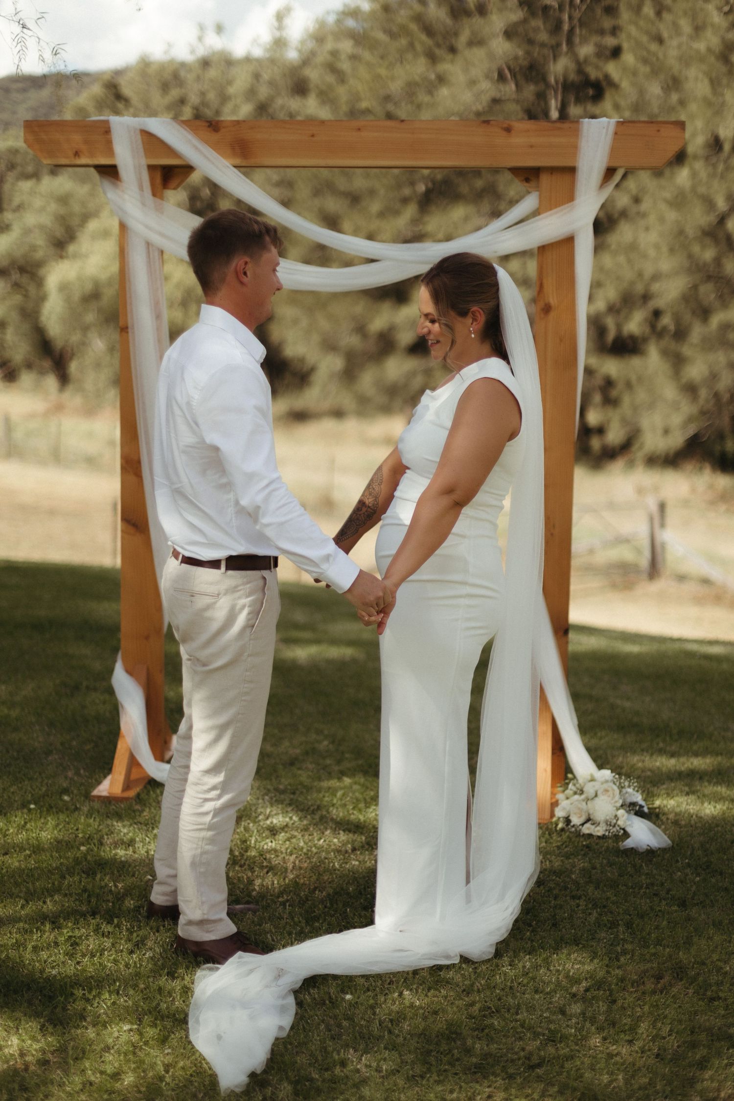 Bride and groom holding hands at outdoor wedding ceremony under rustic wooden arch with white draping.