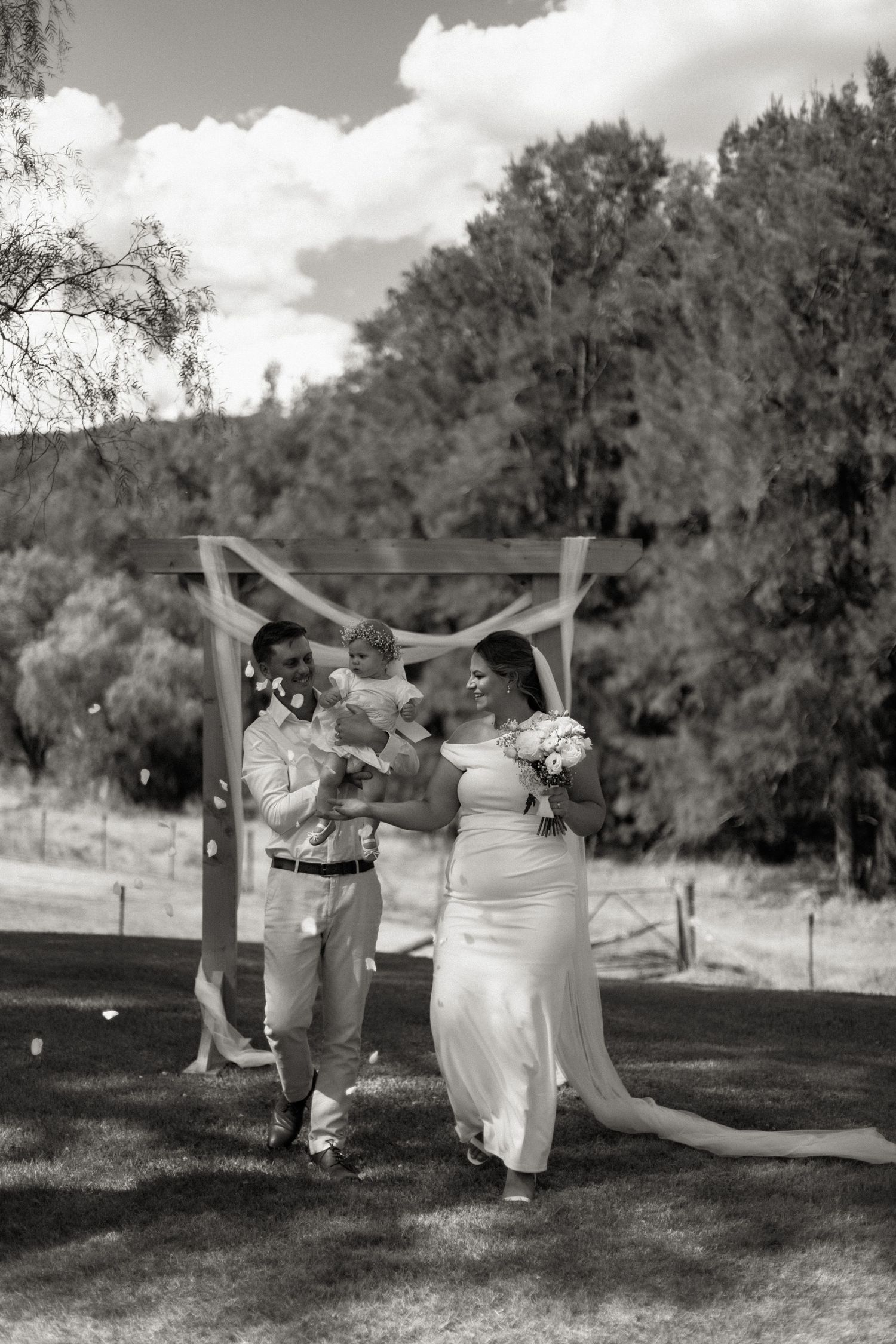 Black and white photo of bride and groom walking with baby after outdoor wedding ceremony under wooden arch.