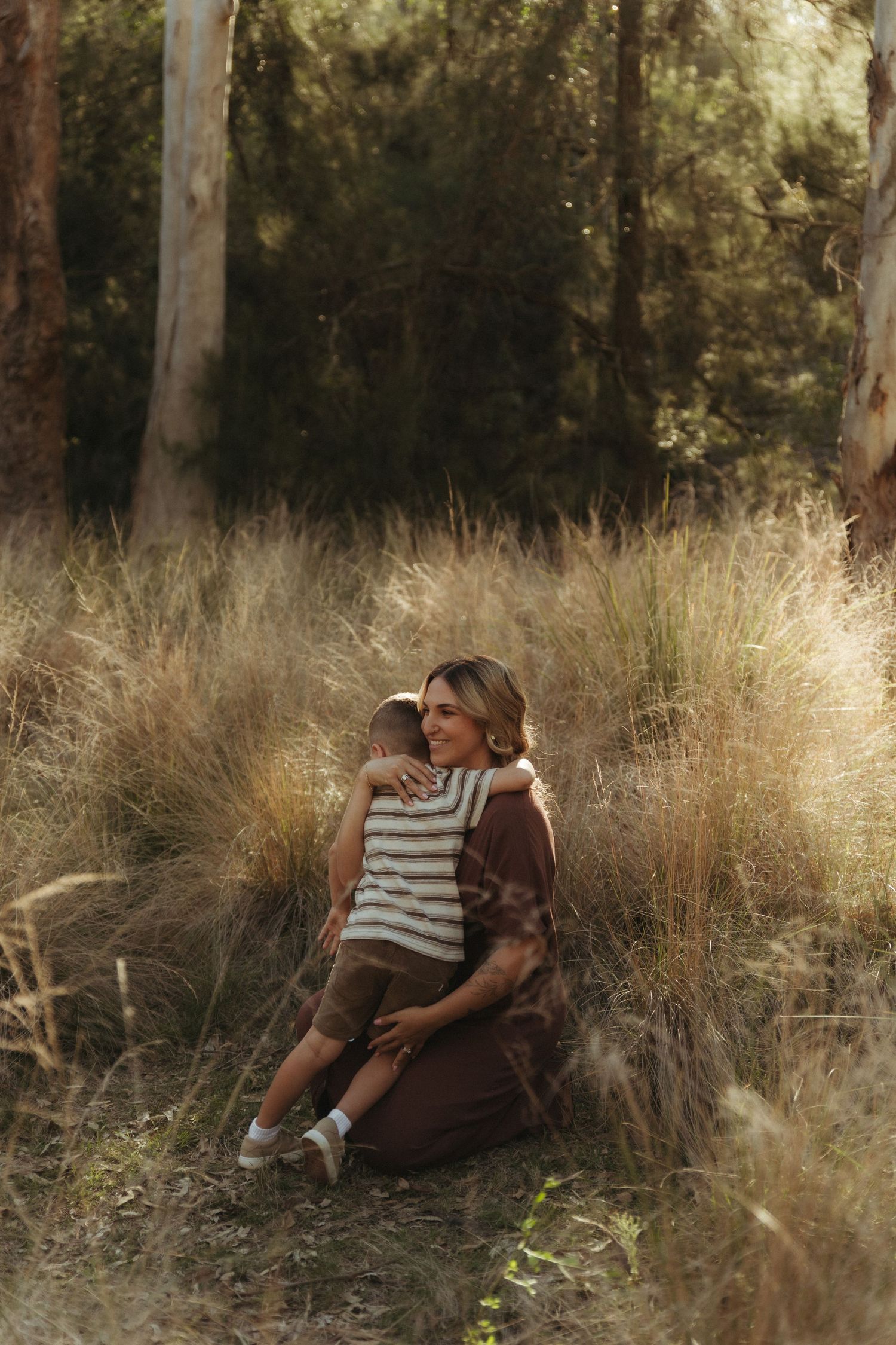 Mother sitting in tall golden grass field hugging her young son in a serene outdoor portrait.