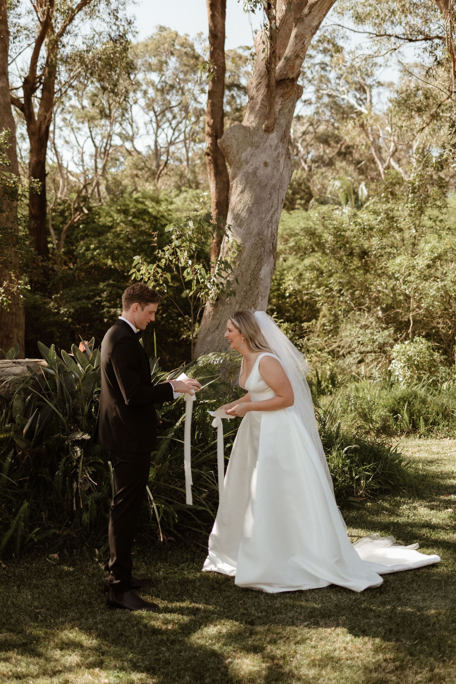 Bride and groom exchange handwritten vows in a sunlit garden surrounded by tall eucalyptus trees.