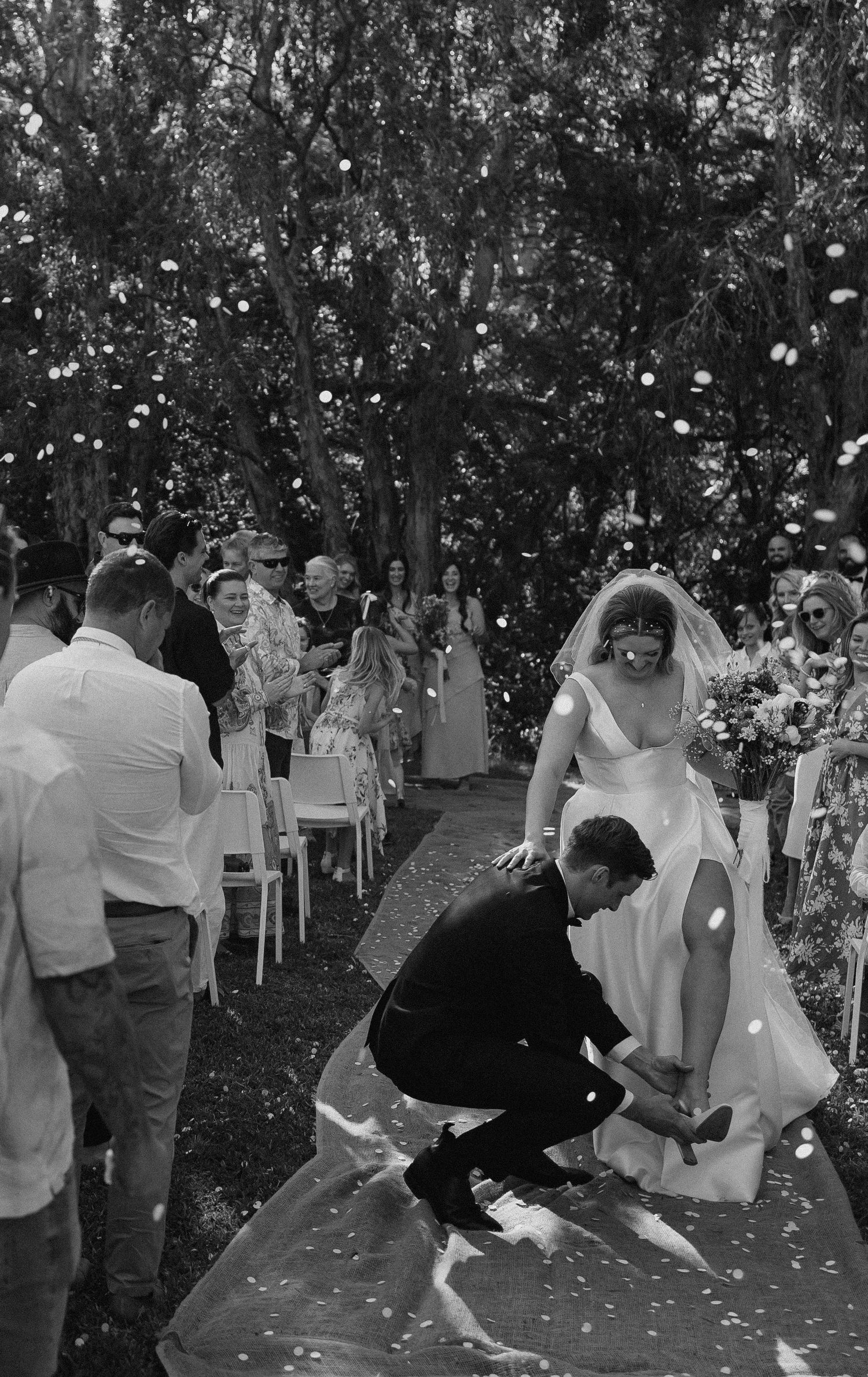 Black and white photo of groom fixing brides shoe on wedding aisle with confetti falling.