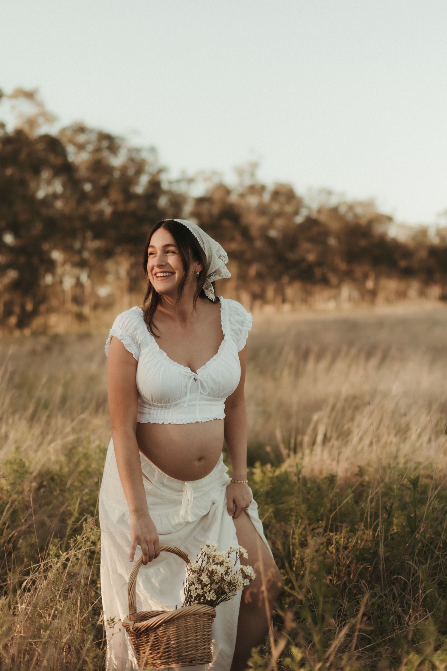 Smiling pregnant woman in white crop top and skirt holds a wicker basket in a golden field.