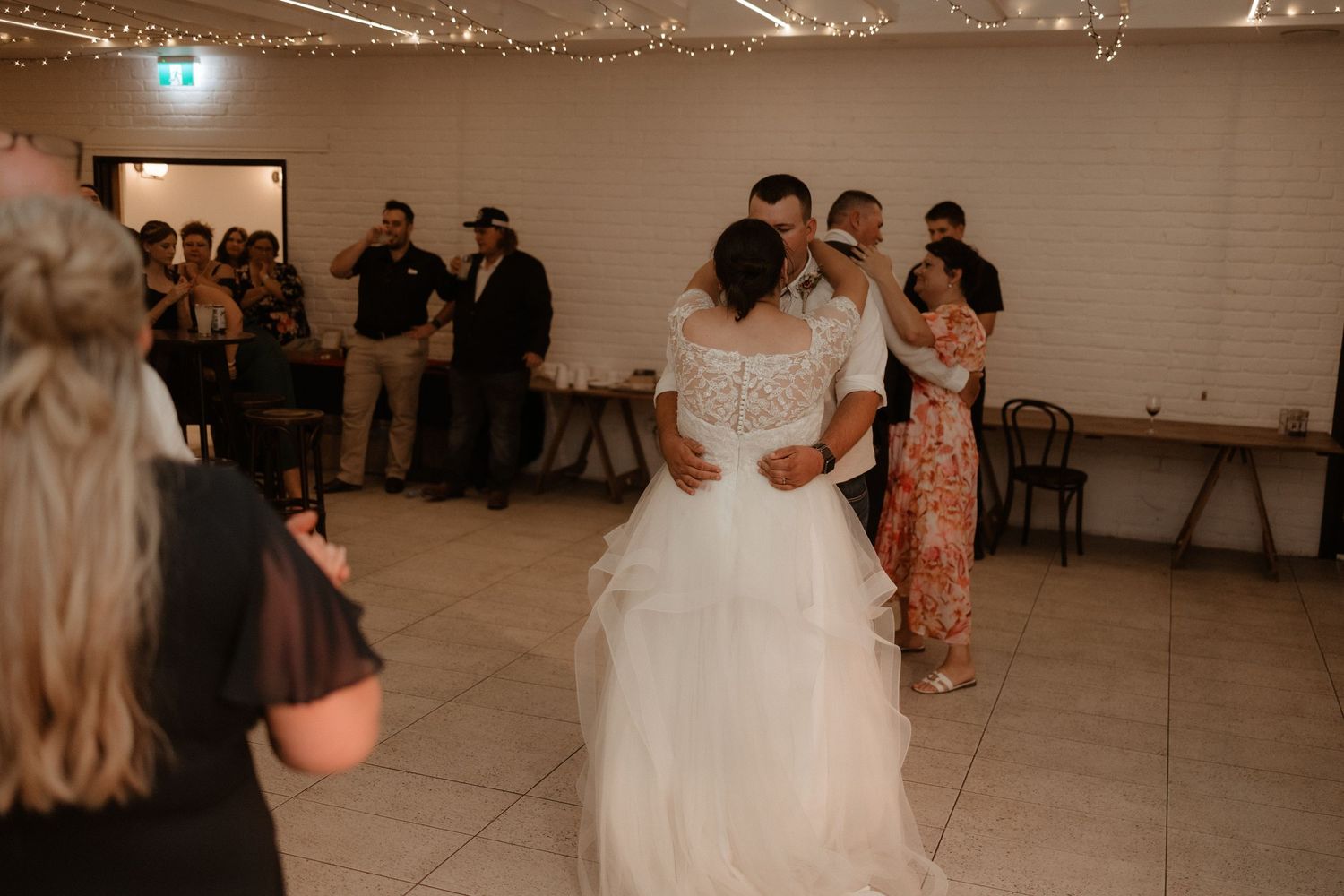 Bride in lace wedding gown dances with groom surrounded by guests at elegant wedding reception.
