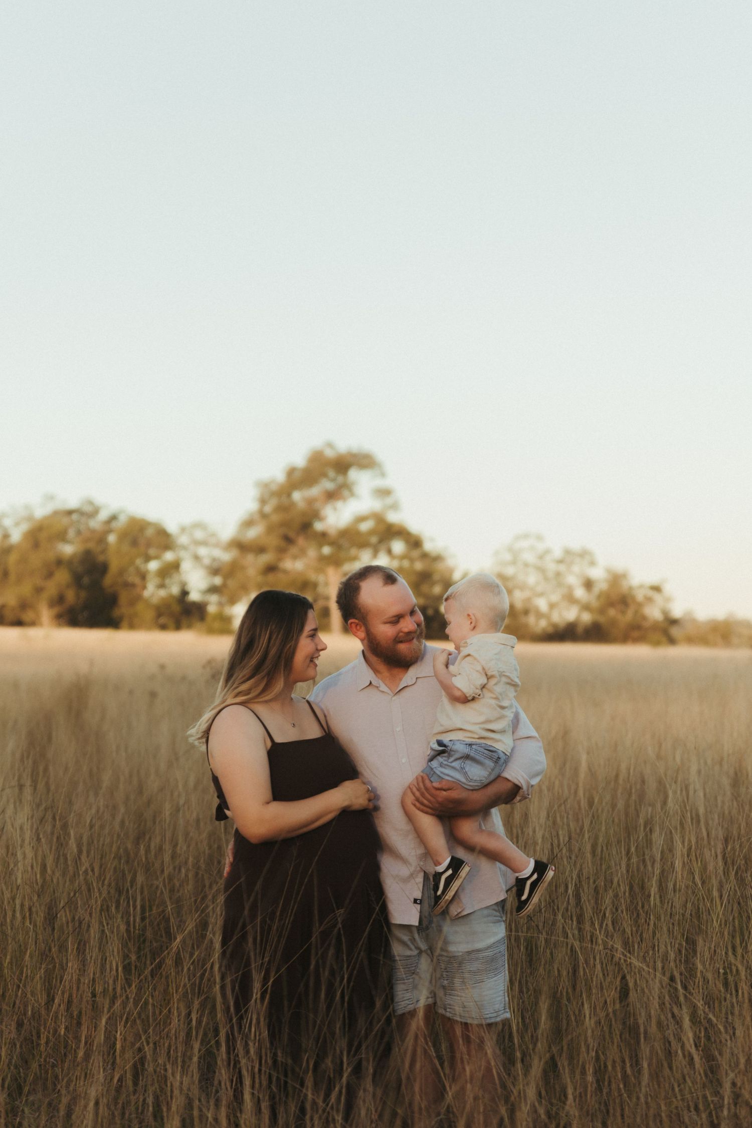 A happy family of three stands in a golden field at sunset, with parents smiling at each other while dad holds their baby.