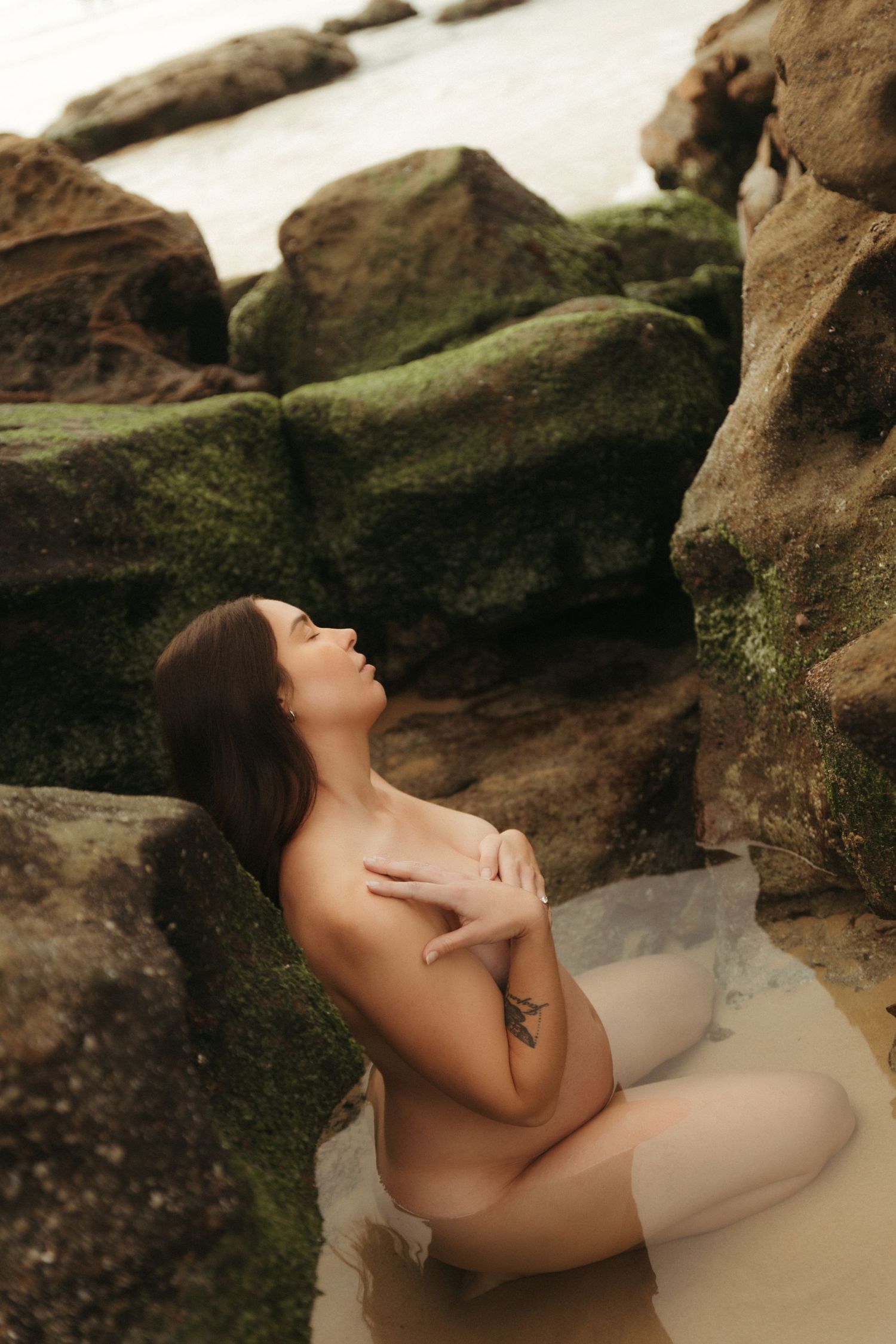 A woman with a tattoo poses gracefully among moss-covered rocks at a beach, with ocean waves in the background.