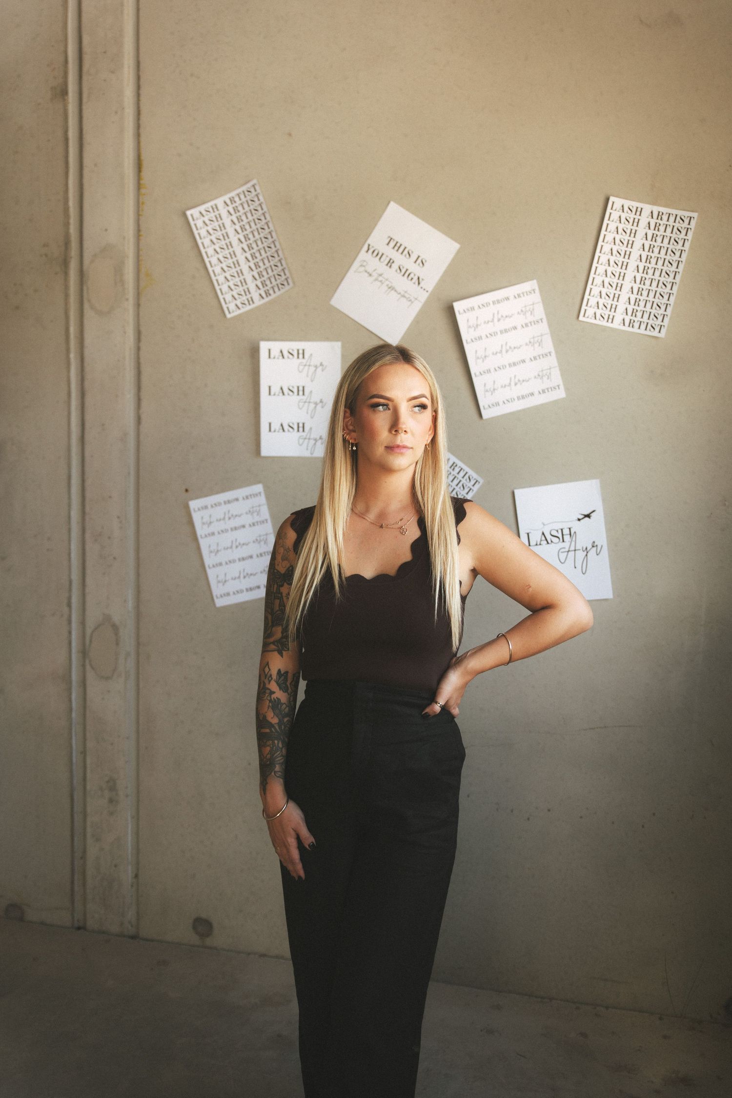 Tattooed blonde woman in black outfit standing before scattered Lash Artist posters on concrete wall.