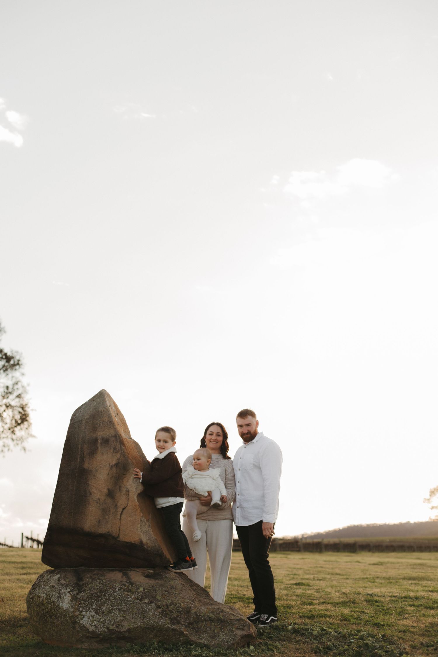 Family of four posing beside large rock sculpture in open field at sunset.