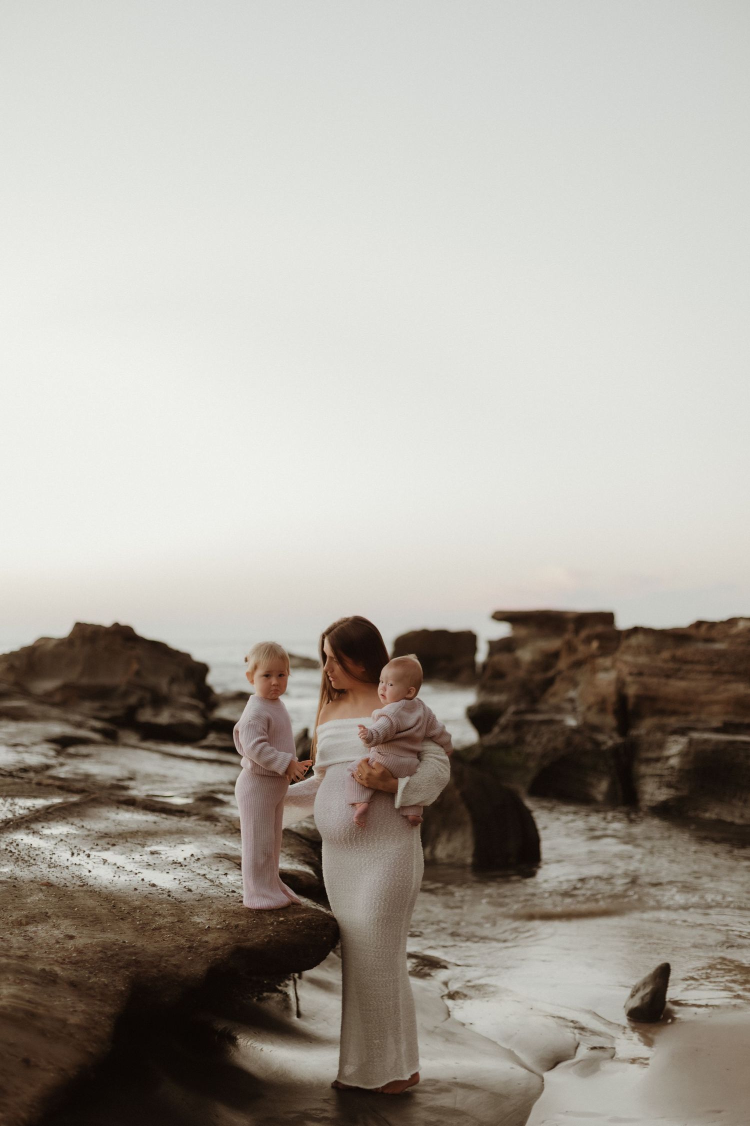Pregnant woman in white gown holding two young children on rocky coastal beach.