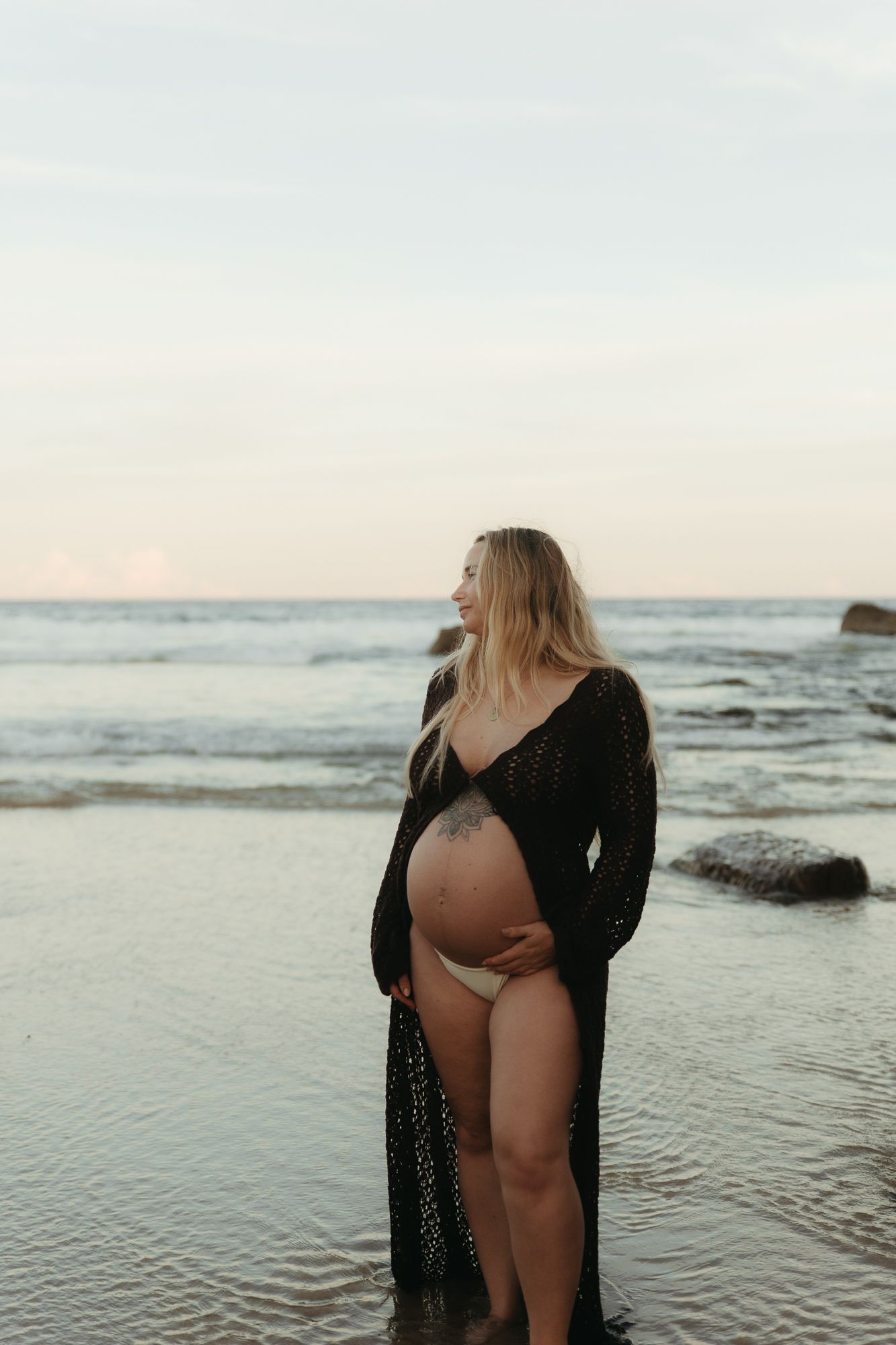 Pregnant woman in black lace cardigan standing on beach at sunset, cradling baby bump, smiling peacefully at ocean waves.