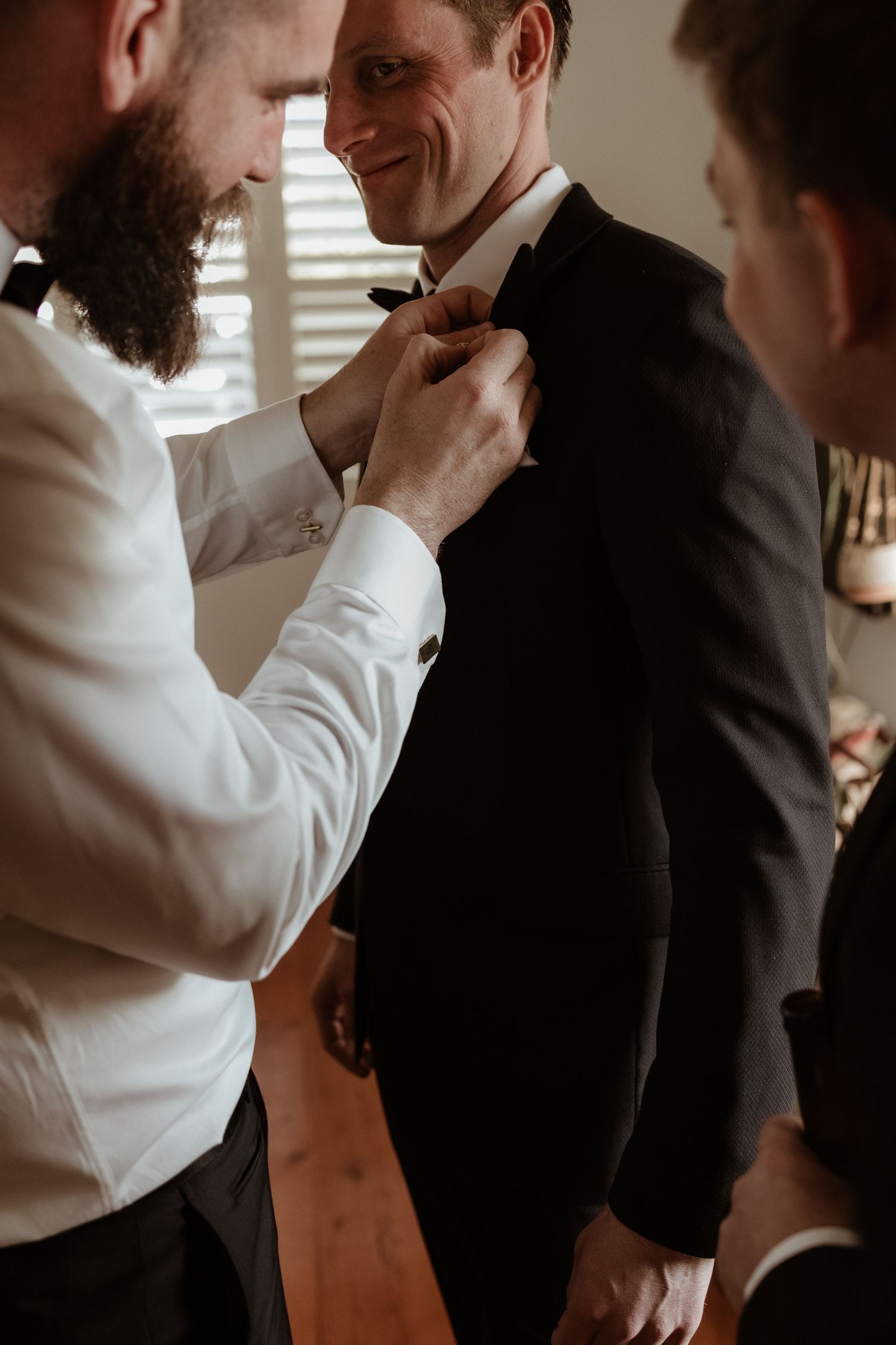 Smiling groom in black tuxedo having his bow tie adjusted by a bearded groomsman.