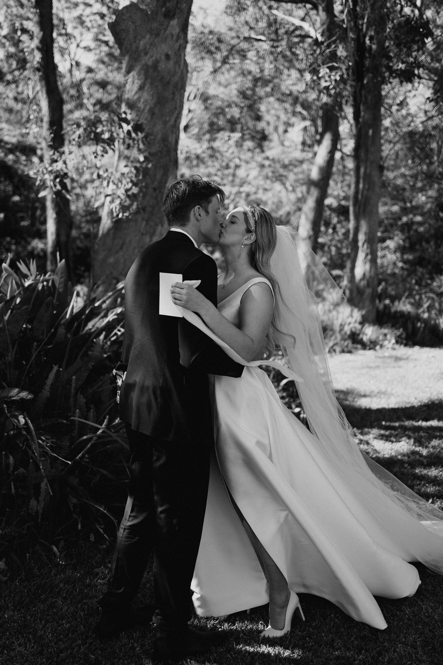 Black and white photo of bride and groom sharing a romantic kiss surrounded by trees on wedding day.