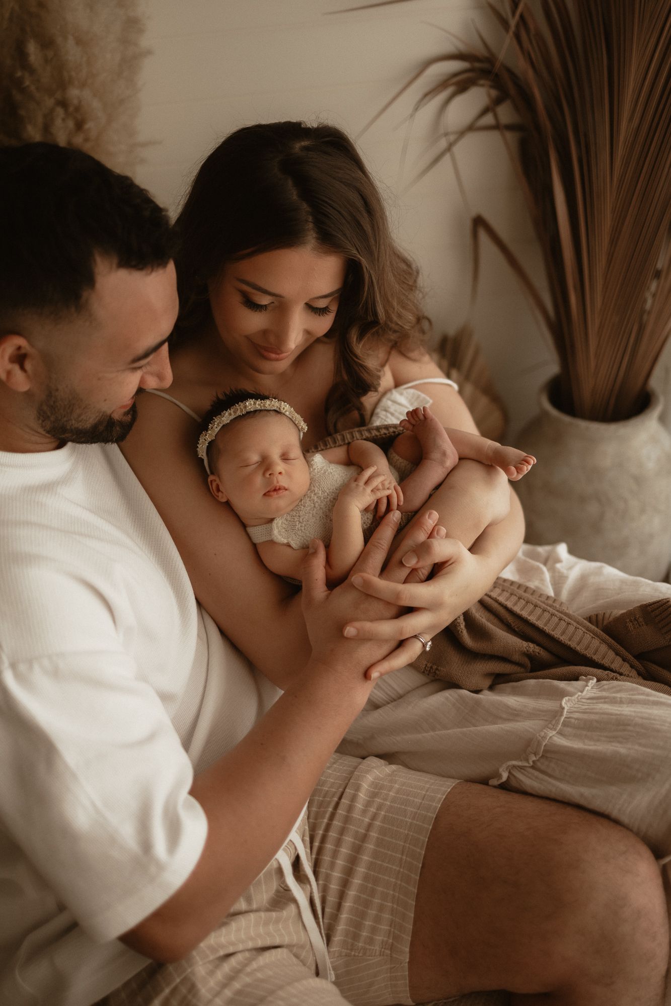 A couple tenderly holds their sleeping newborn baby, dressed in a cream outfit with a headband, in a warm boho-styled room.