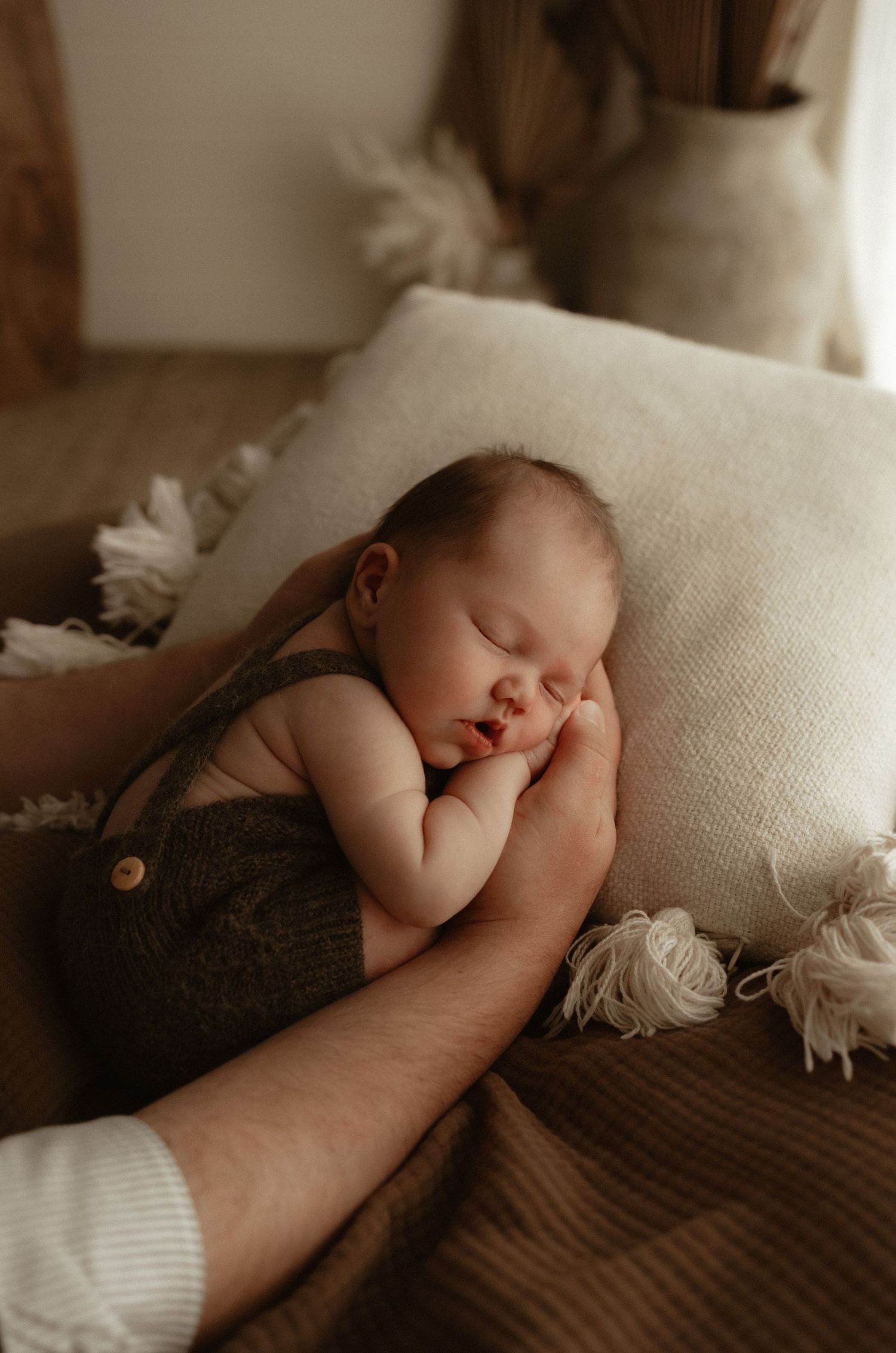 Newborn baby sleeping peacefully in brown romper, cradled in adult arms on cream textured pillow.