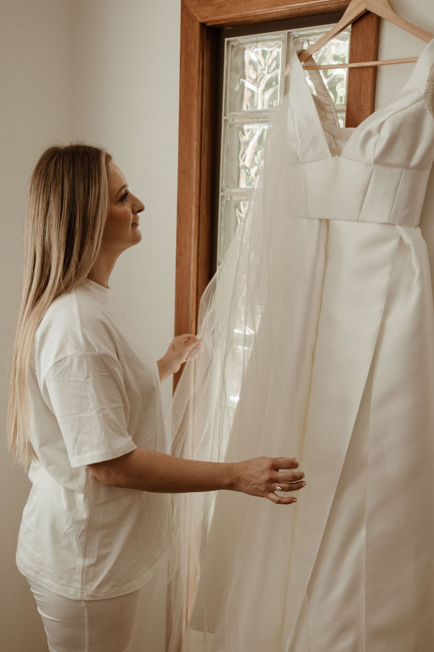 Smiling blonde bride in white t-shirt admiring her wedding dress hanging in a doorway.