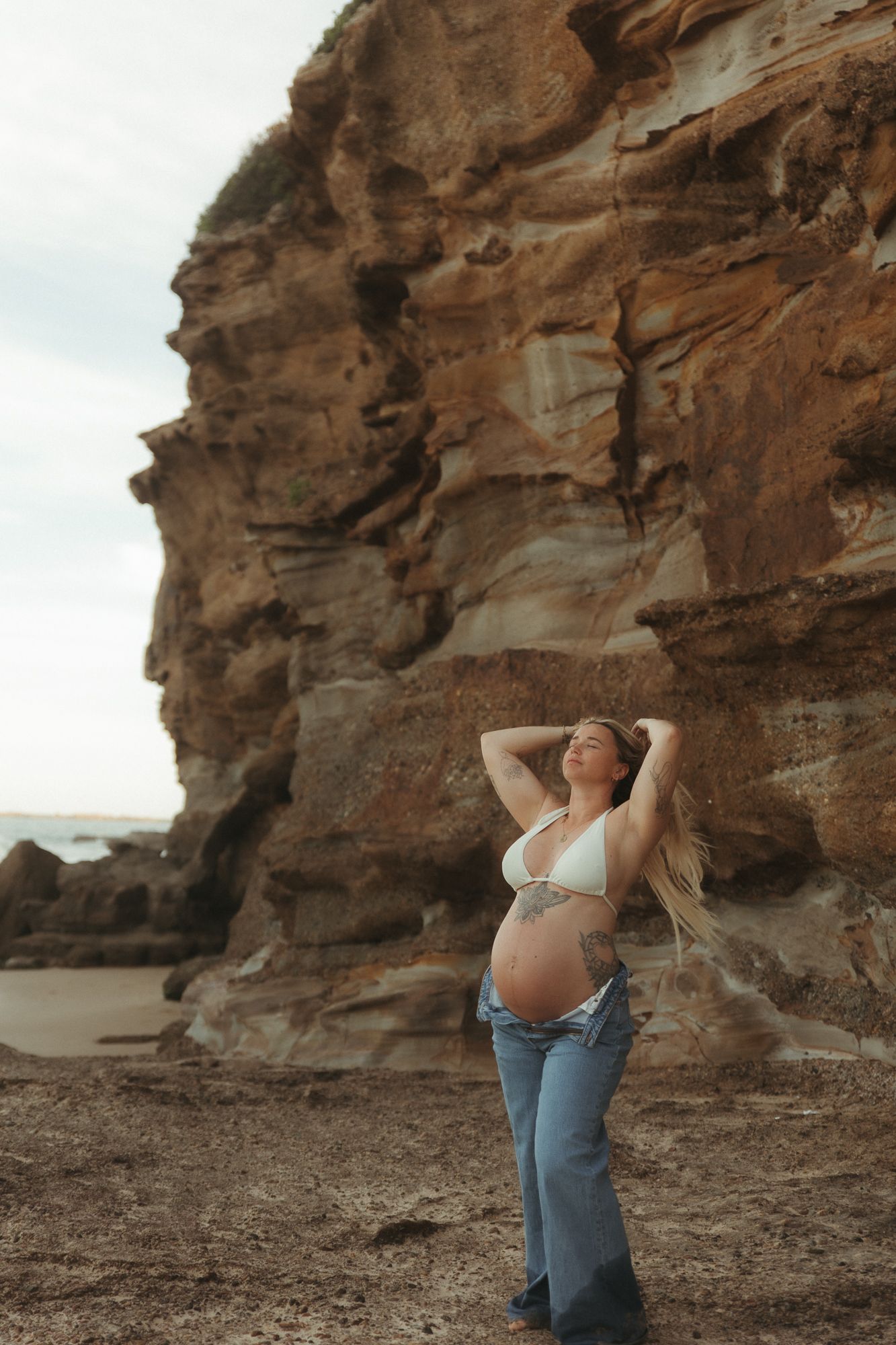 Pregnant woman in white bikini top and jeans posing against rugged sandstone cliffs at a rocky beach.