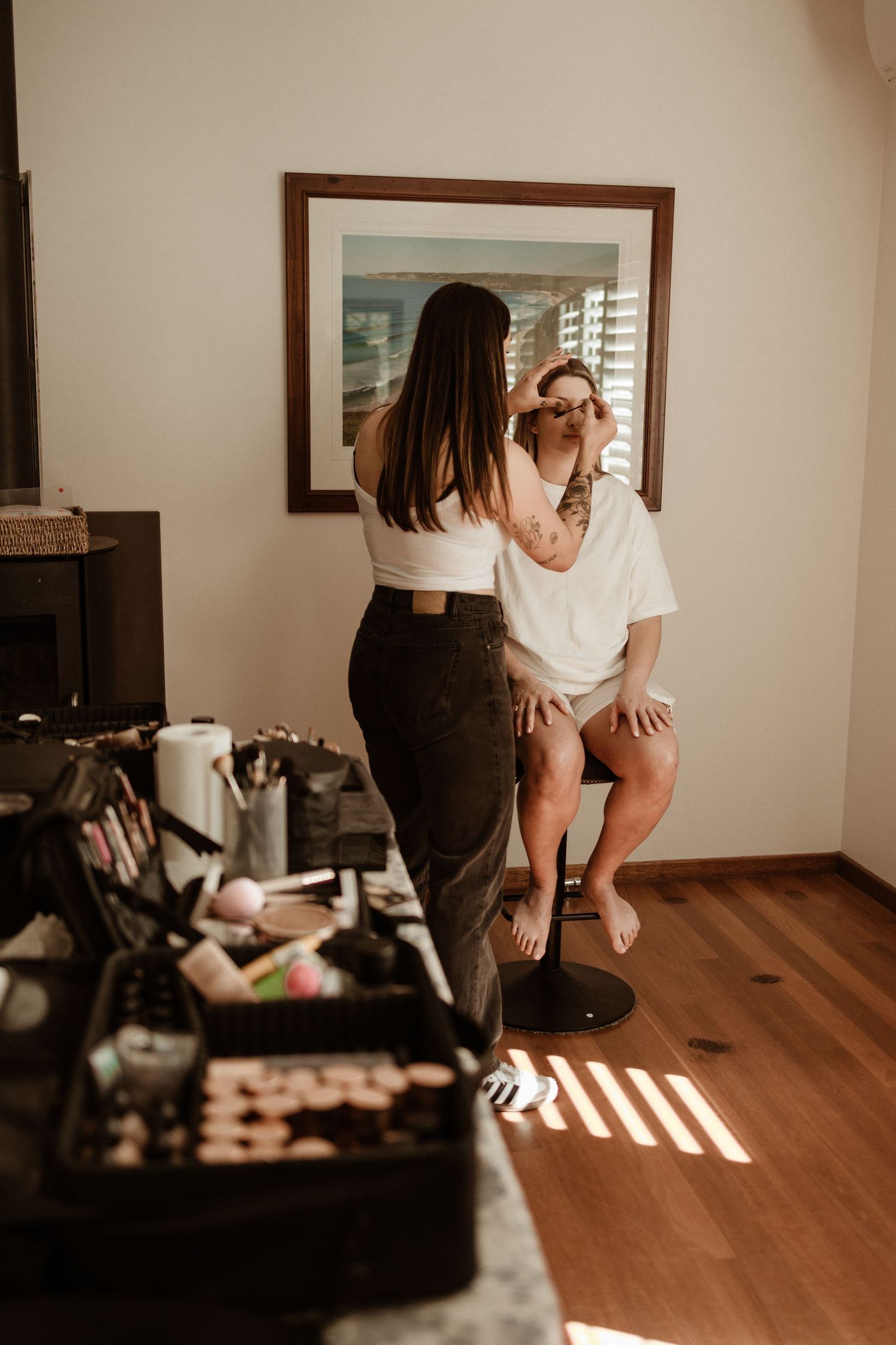 Makeup artist applying makeup to a bride-to-be seated before a mirror in a sunlit hotel room.