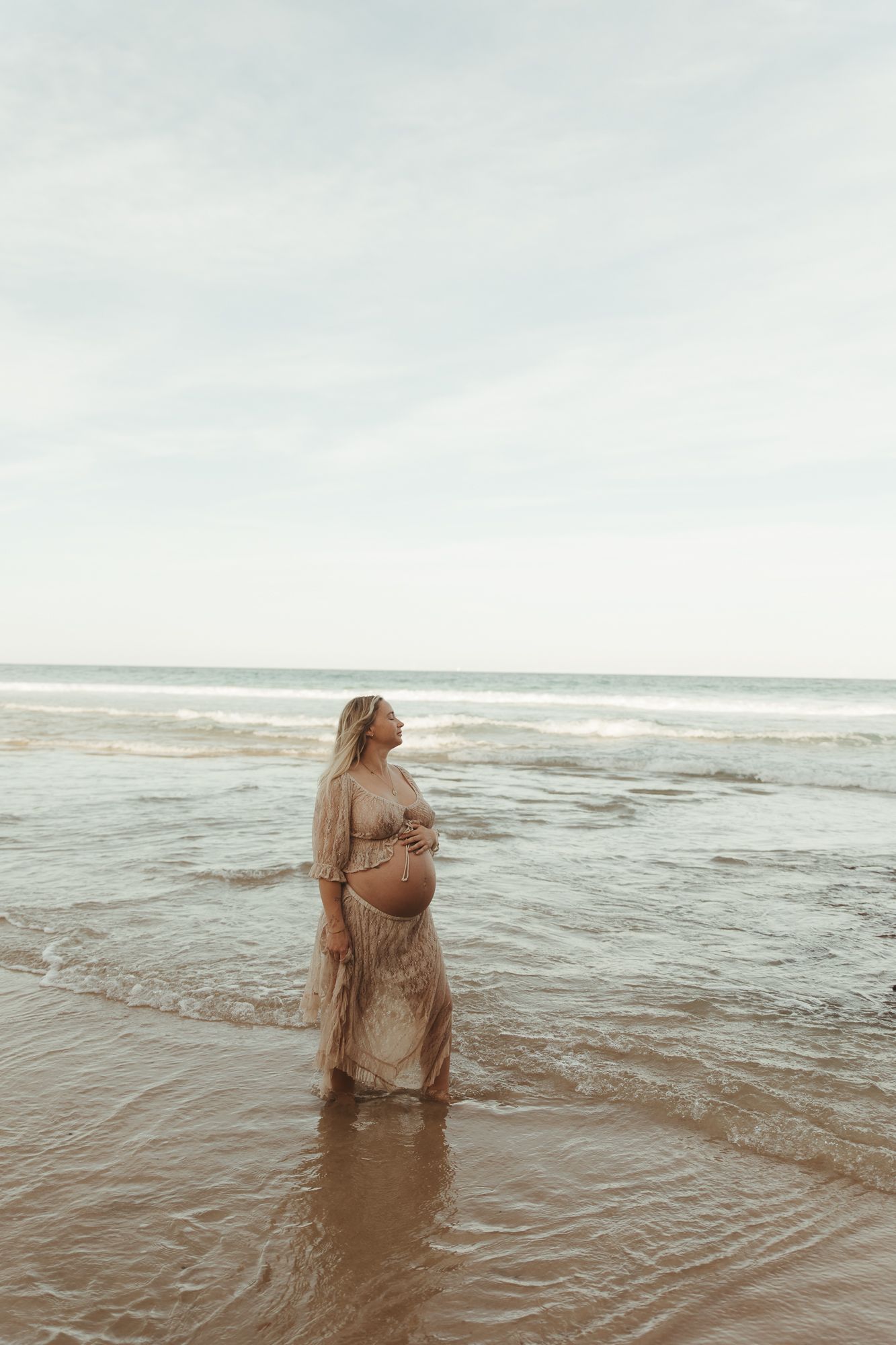 Pregnant woman in sheer beige dress standing in ocean waves on a beach, cradling her baby bump.