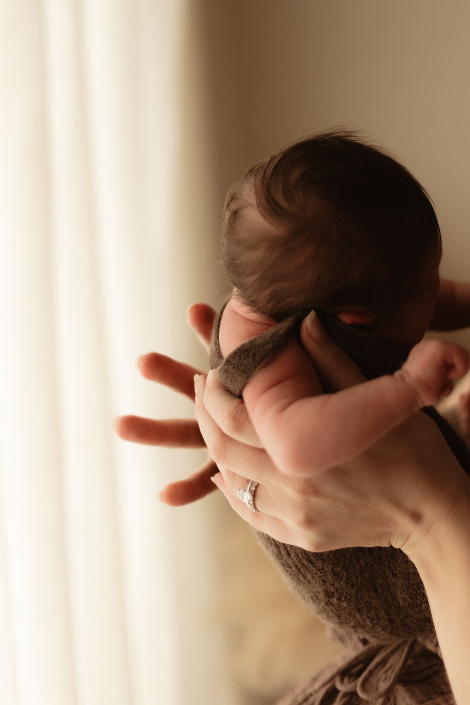 A newborn baby with dark hair being cradled in an adult's hands, tiny fingers spread wide near a bright window.