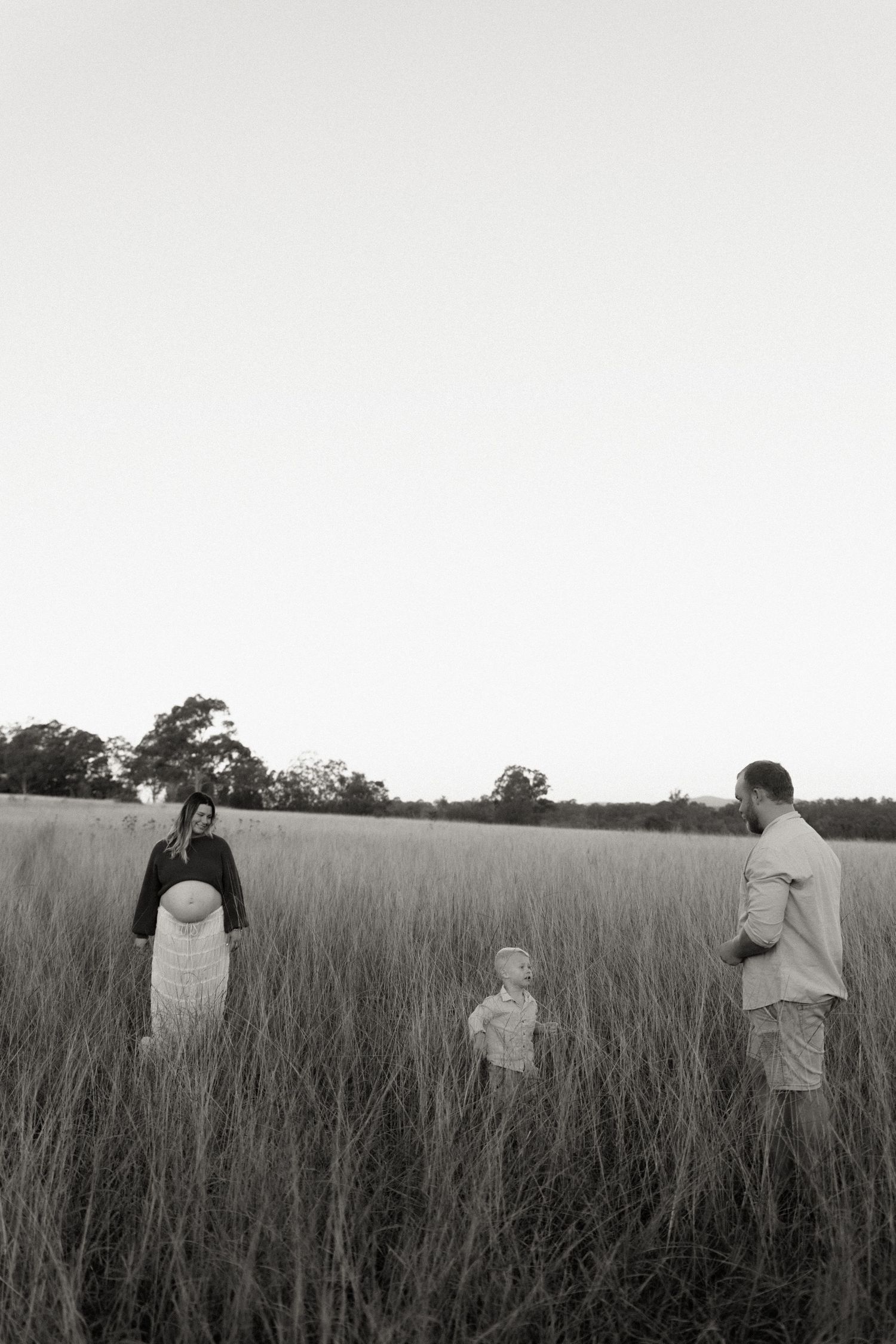 Black and white photo of a family with a pregnant woman, child, and man standing in a tall grass field at dusk.