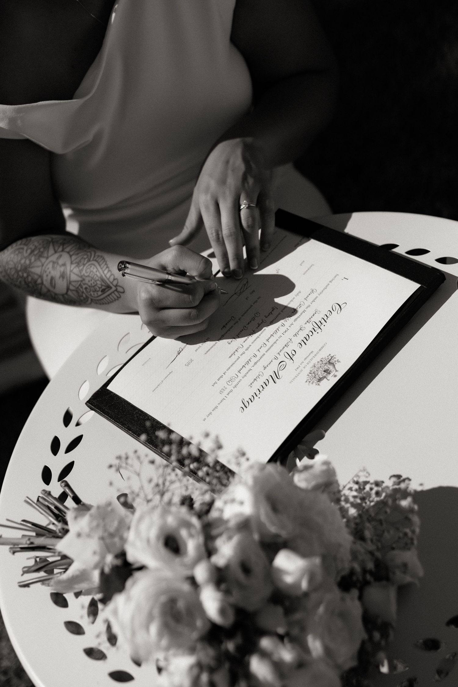 Black and white close-up of couple signing marriage certificate with wedding bouquet nearby.