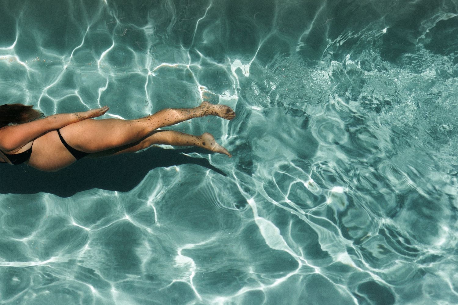 Aerial view of a woman in a black bikini swimming through clear turquoise pool water.