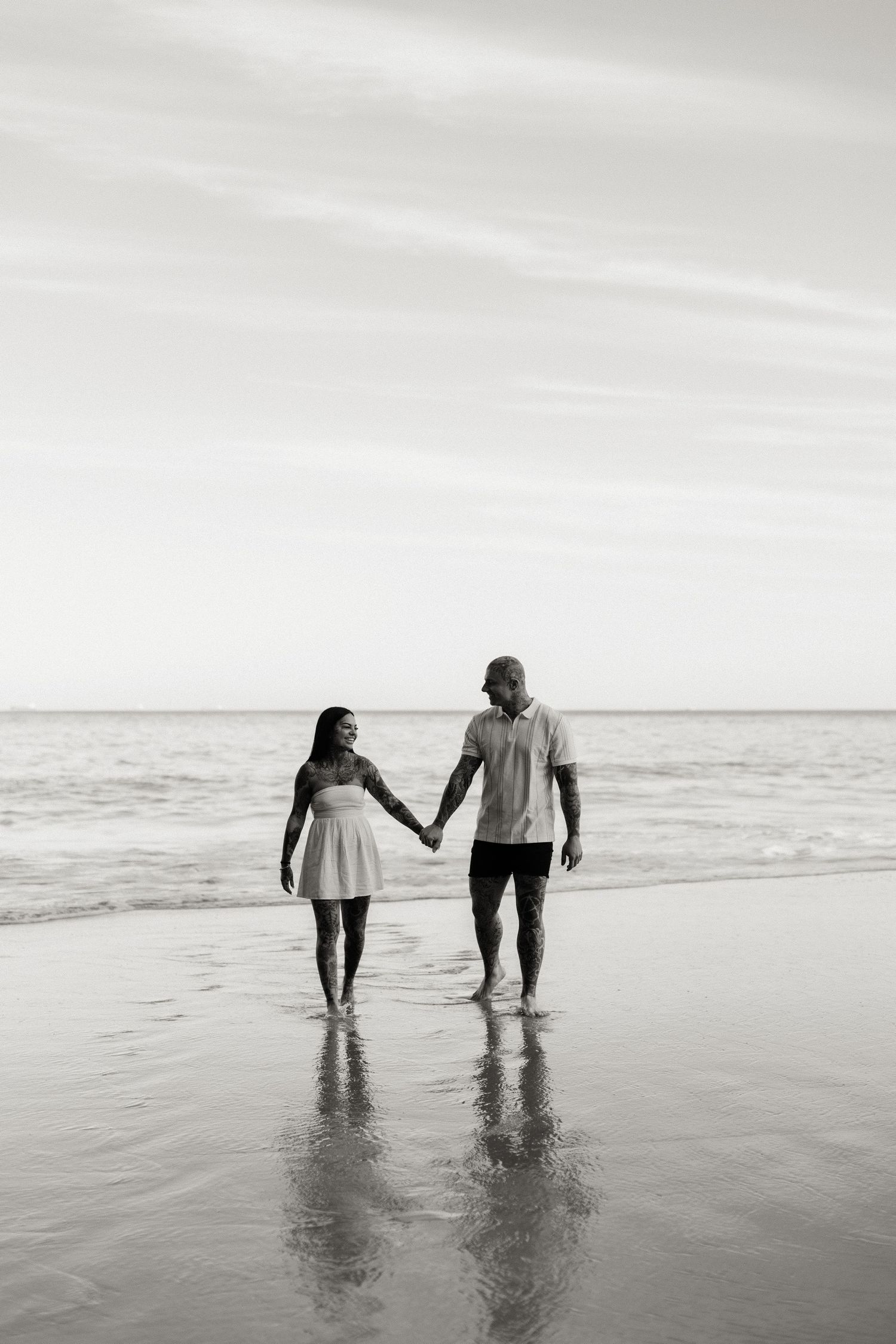 Black and white photo of a couple holding hands while walking barefoot on a beach with ocean waves in the background.