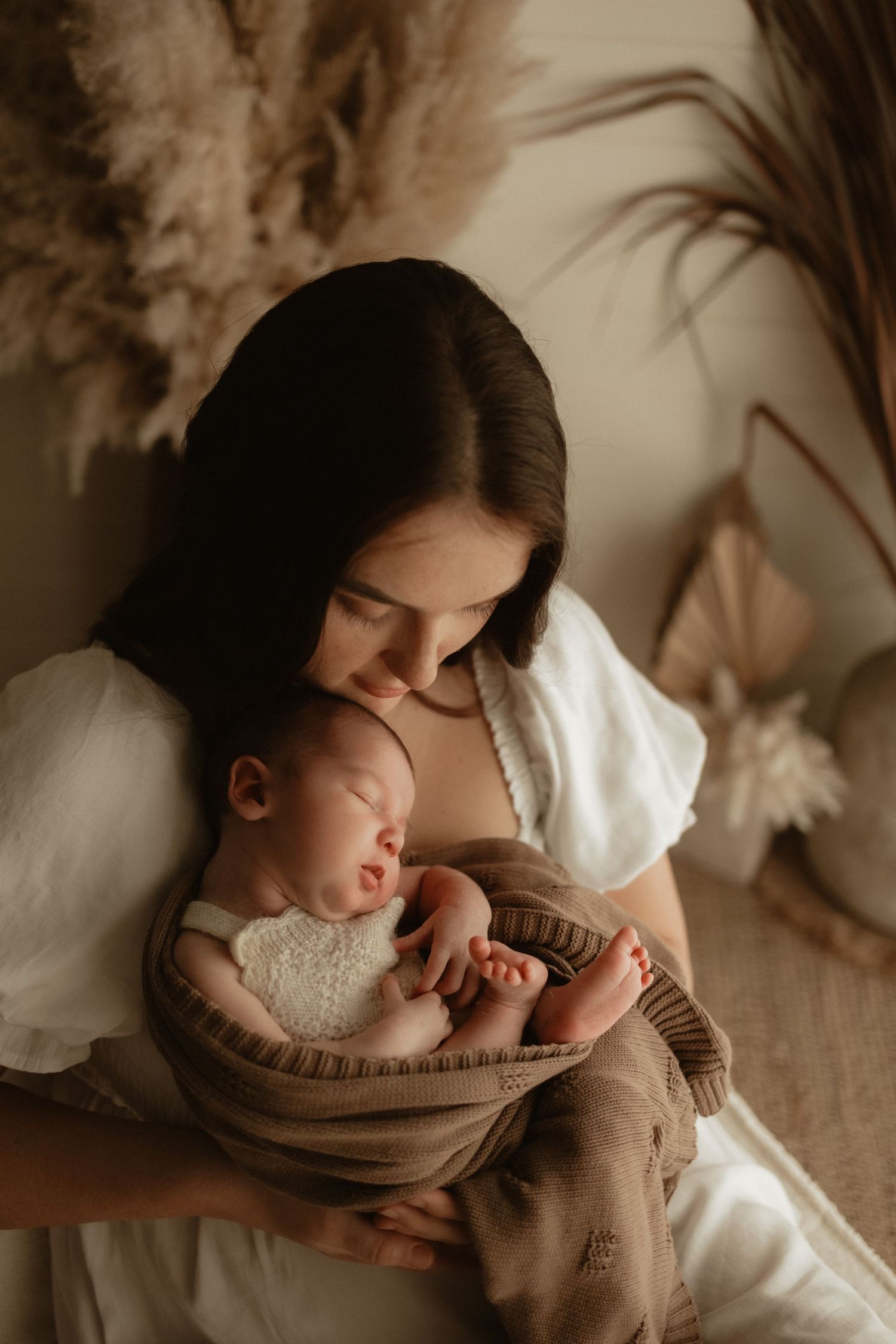 Mother in white dress lovingly gazing at her sleeping newborn wrapped in a brown knit blanket.