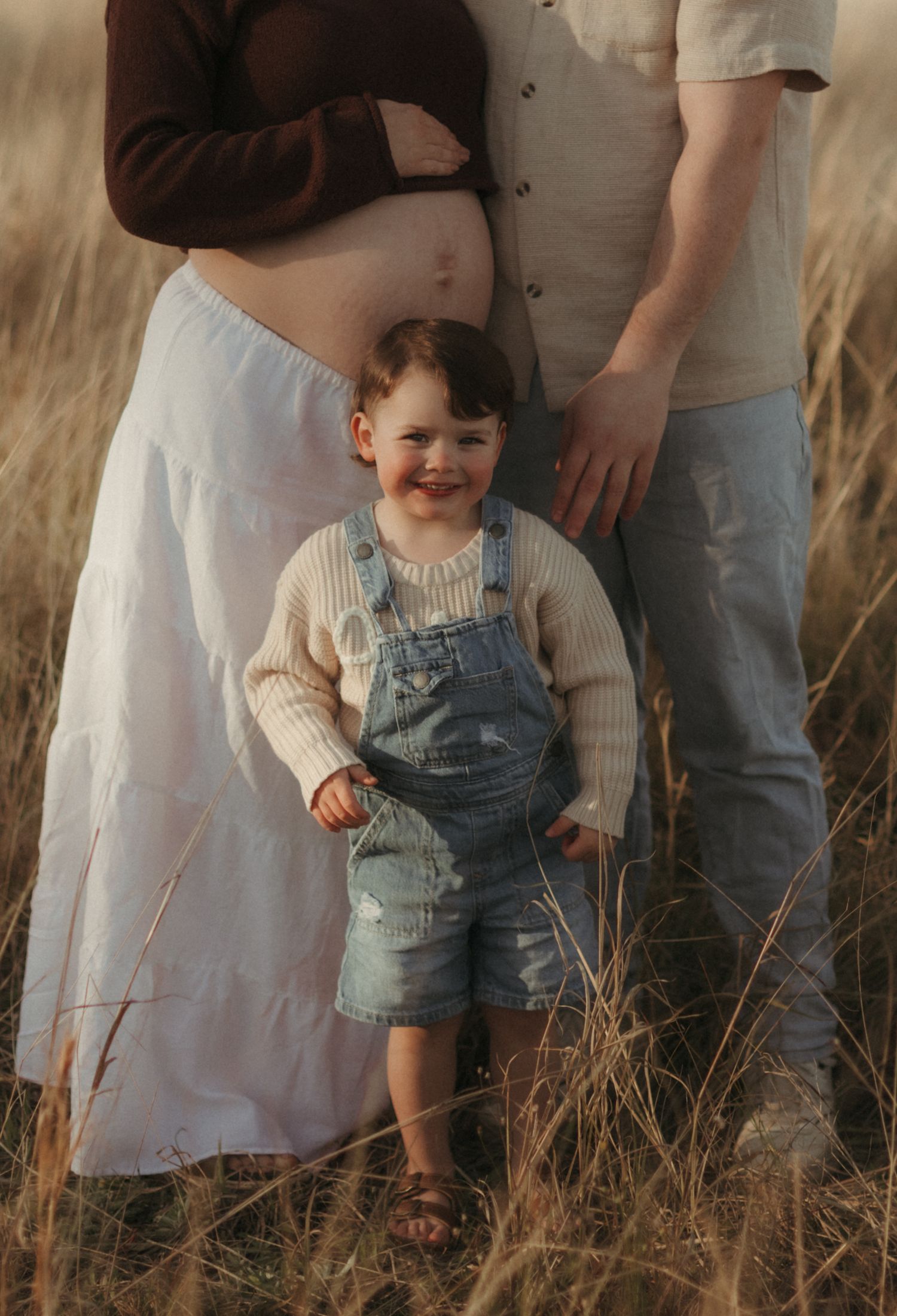 Toddler in denim overalls smiling between pregnant mother and father in golden grass field.