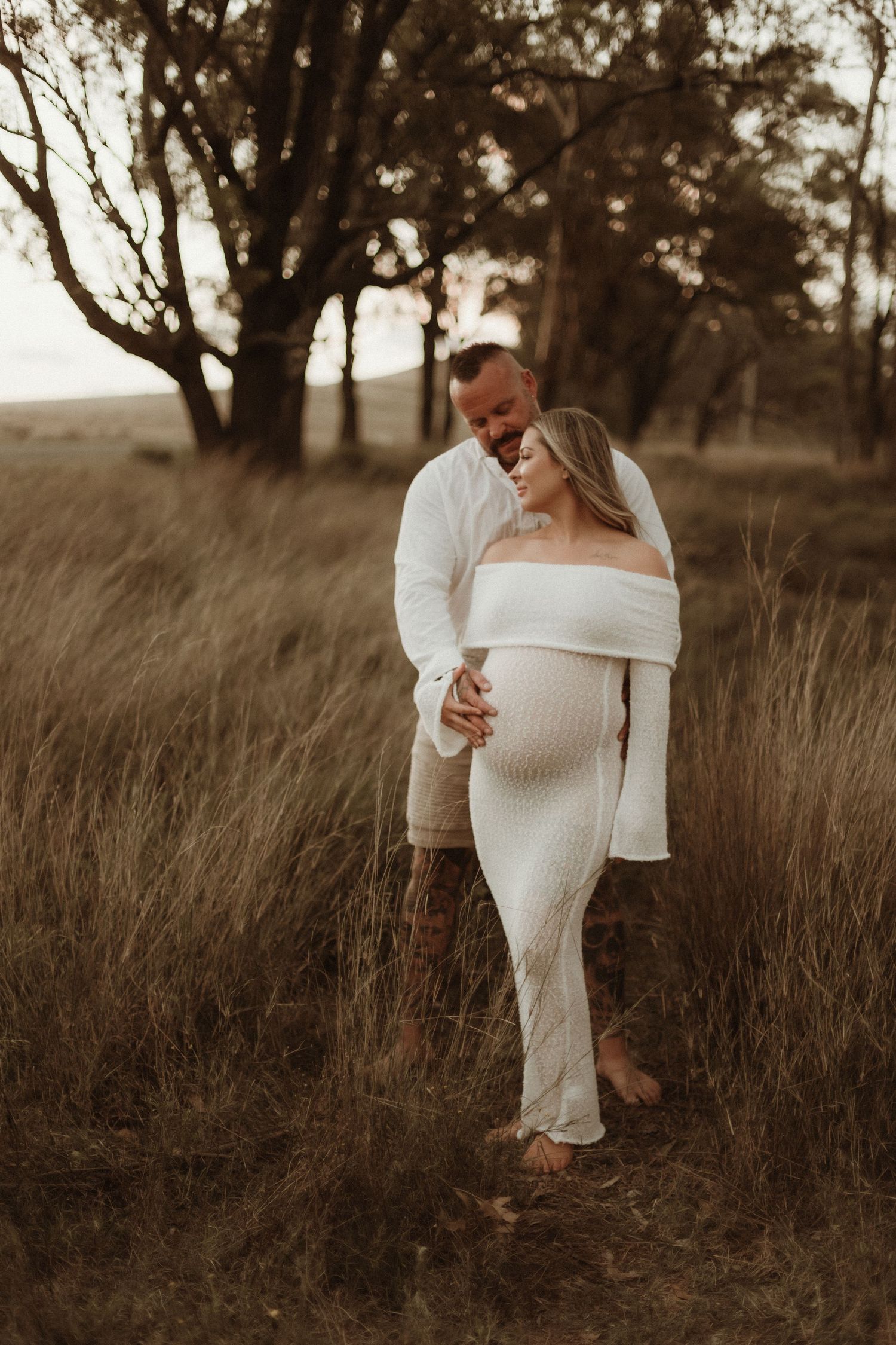 Couple sharing tender moment in golden tall grass field at sunset, woman in white maternity dress.
