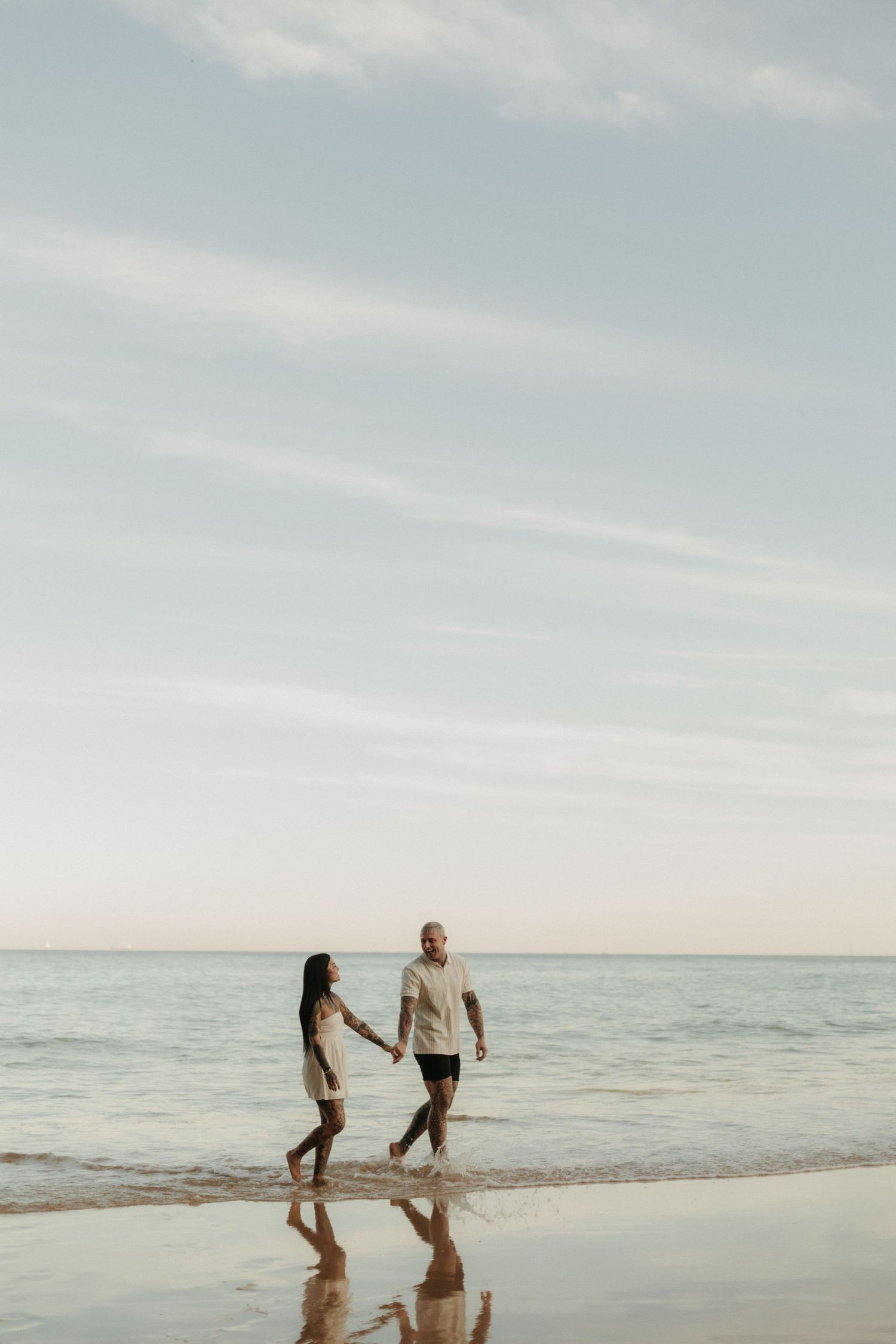 Couple holding hands walking along calm beach shoreline at sunset with reflections.