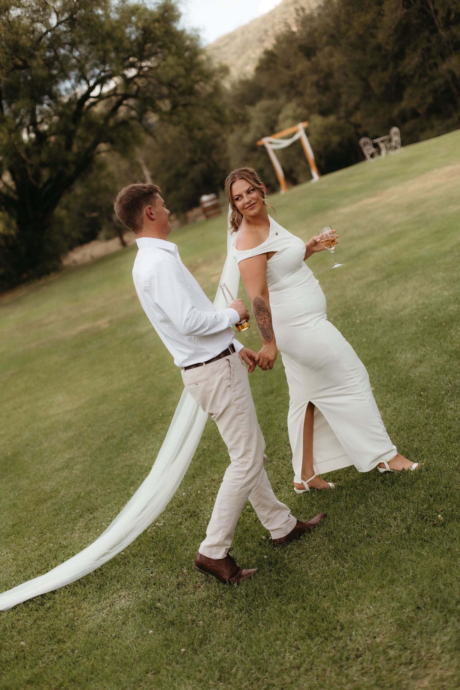 Smiling bride and groom walk together on green lawn with flowing veil and champagne.