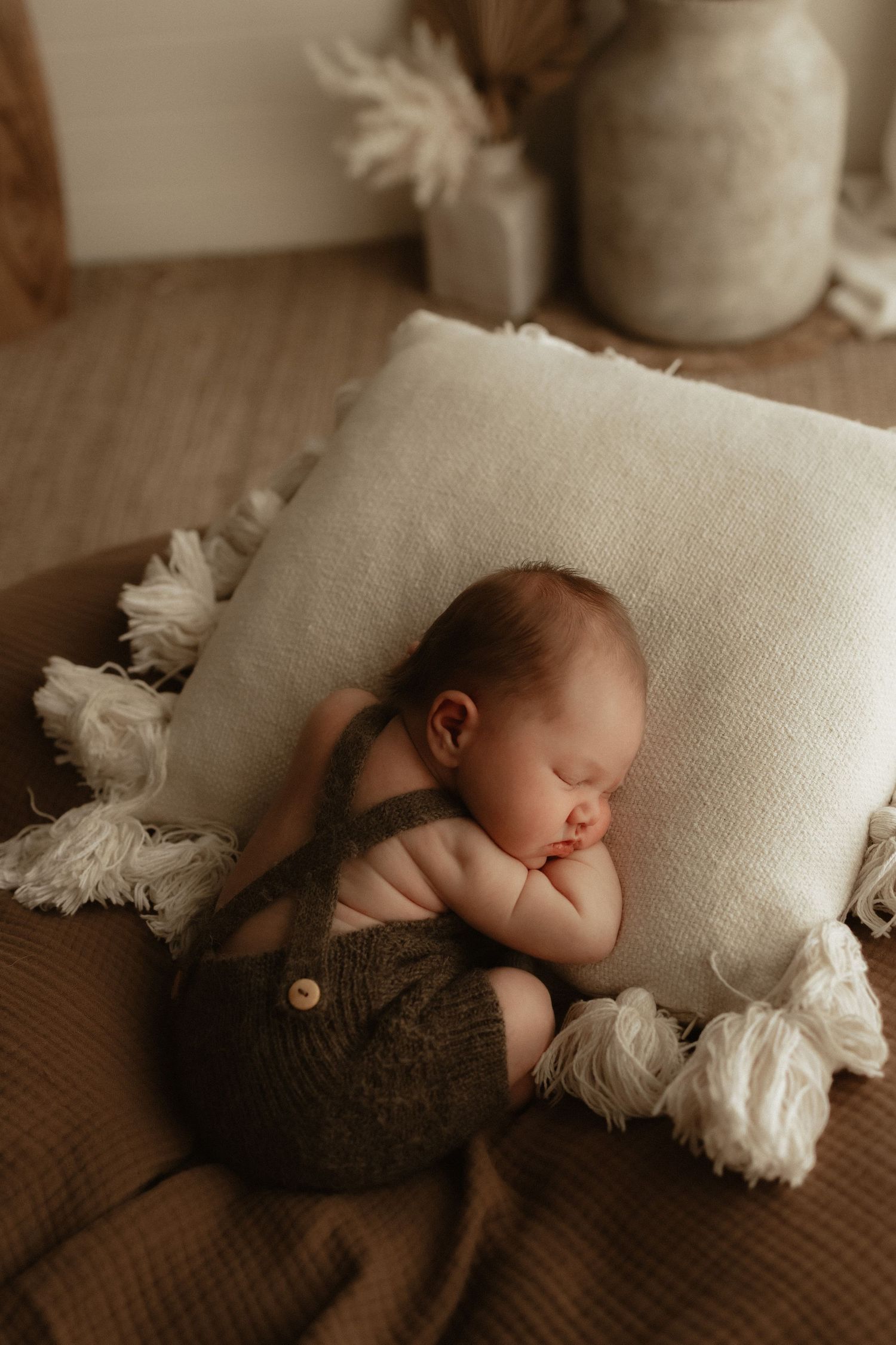 Sleeping newborn baby in olive knit overalls curled on a cream tasseled pillow in a boho nursery setting.
