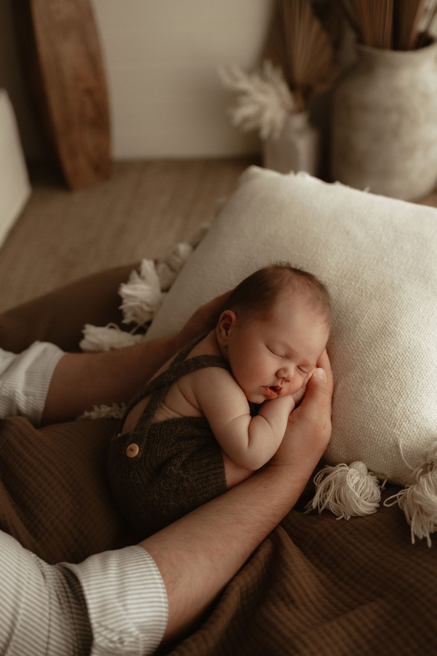 Sleeping newborn baby in olive romper resting on parent's arm surrounded by boho decor.