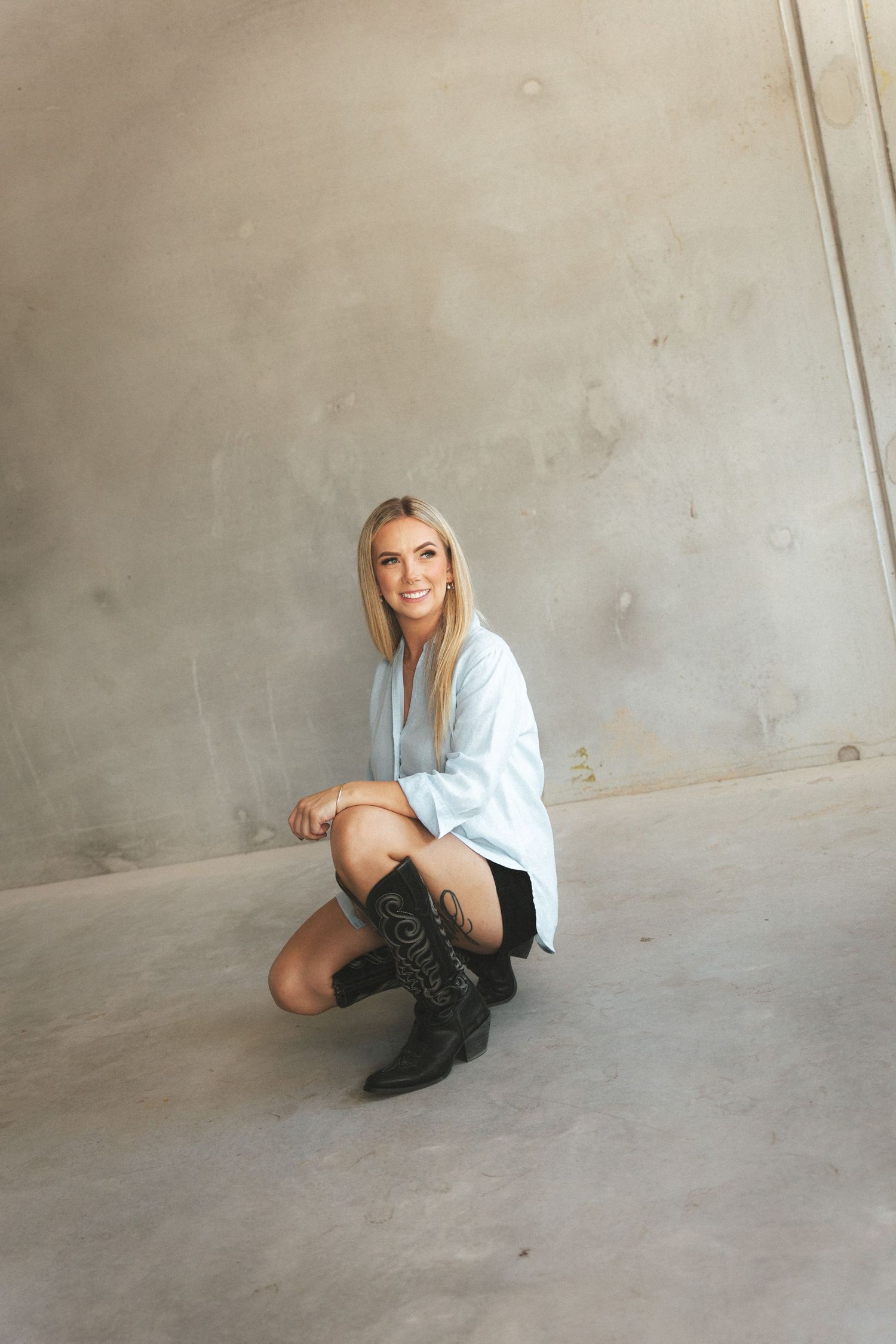 Smiling blonde woman crouching in light blue shirt, black shorts, and cowboy boots against concrete wall.
