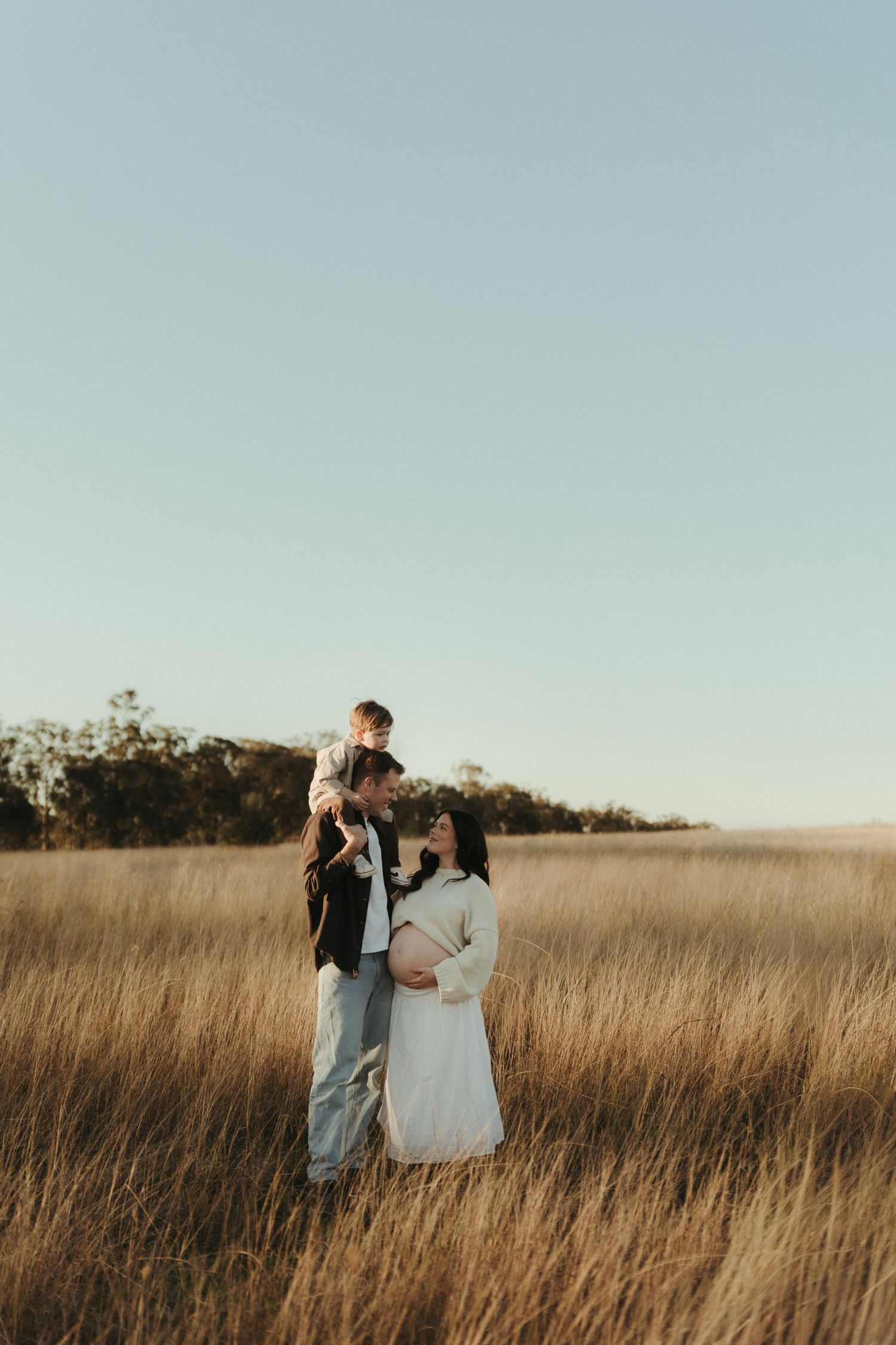 Pregnant woman in white dress with partner holding toddler in golden grass field during maternity photoshoot.